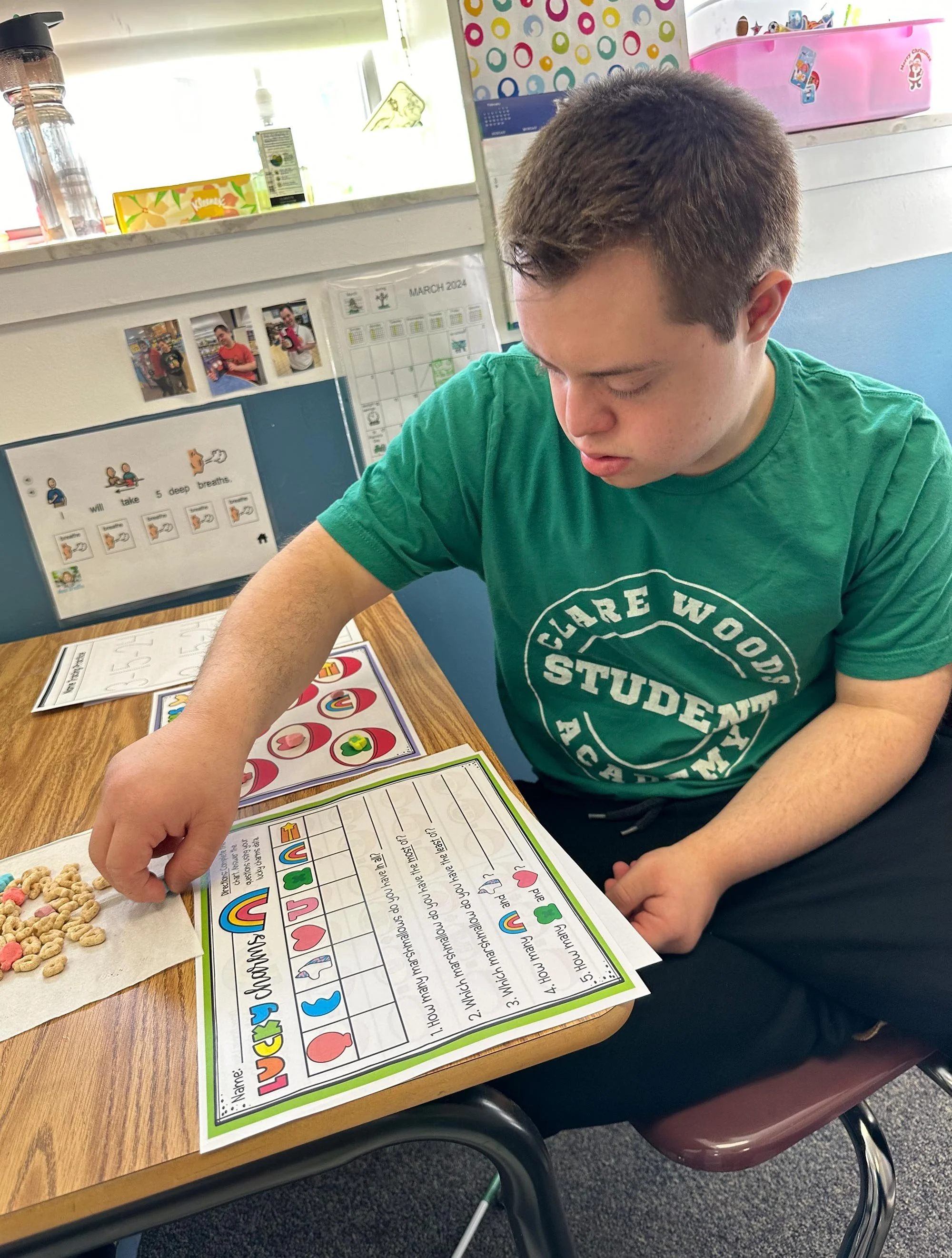A young boy in a green T-shirt sitting at a wooden table with educational worksheets and snacks, participating in a learning activity, in a classroom setting with colorful posters and decorations in the background.