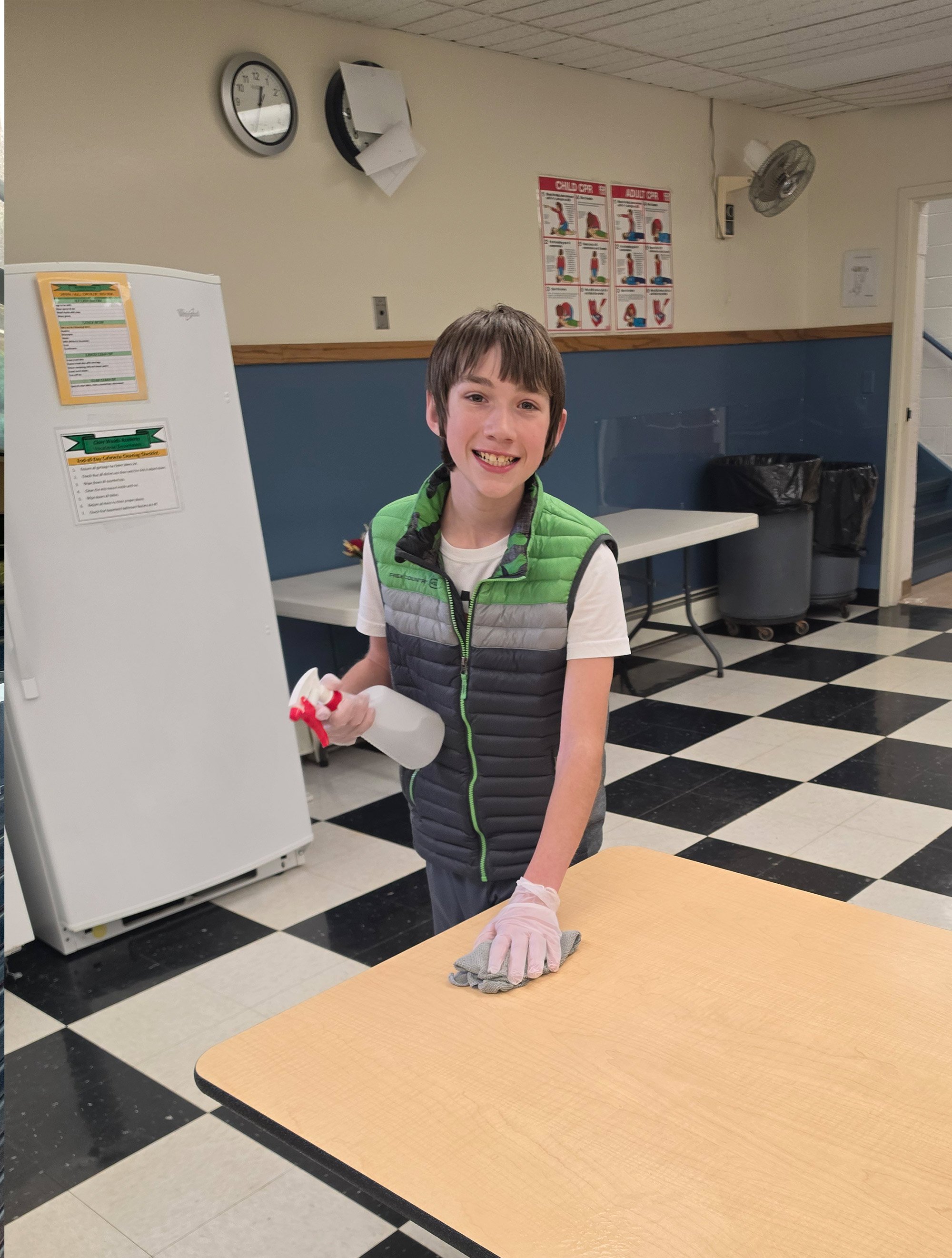 A young boy smiling, wearing a green and gray vest, cleaning a table with a gray cloth in a room with black and white checkered floor.