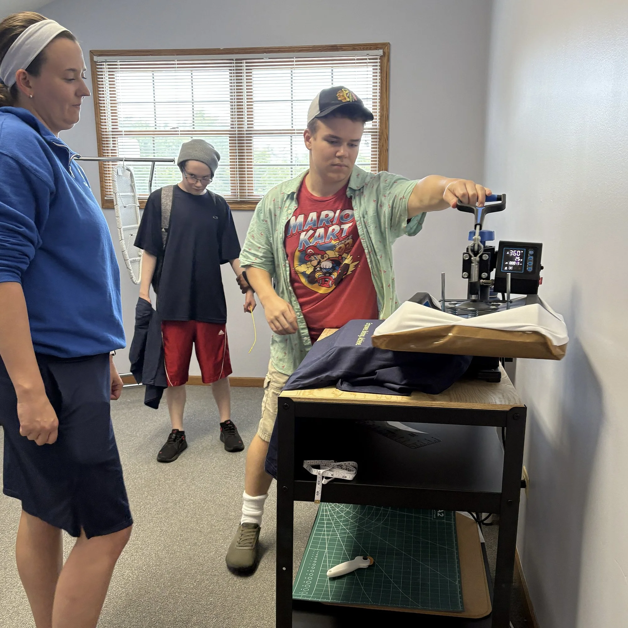 Three day program participants in a room, observing and operating a machine on a table to heat-press t-shirts. One person is operating the machine, another with a backpack observes, and a supervisor watches attentively.