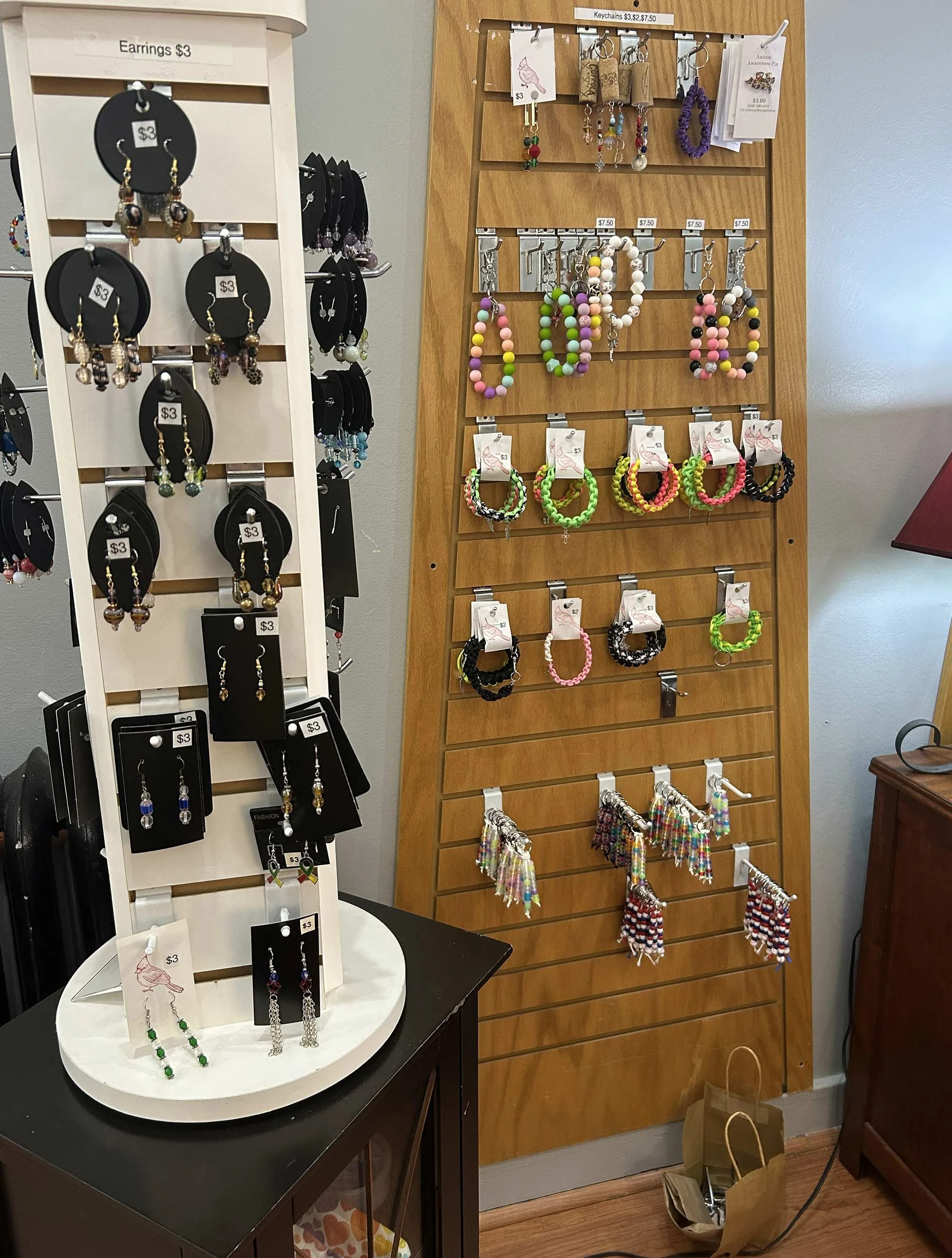 A display of earrings, necklaces, and bracelets on a rotating stand and wall hooks in a store. The display includes colorful beaded jewelry, earrings, and pins, with price tags indicating $3 and $7.50.