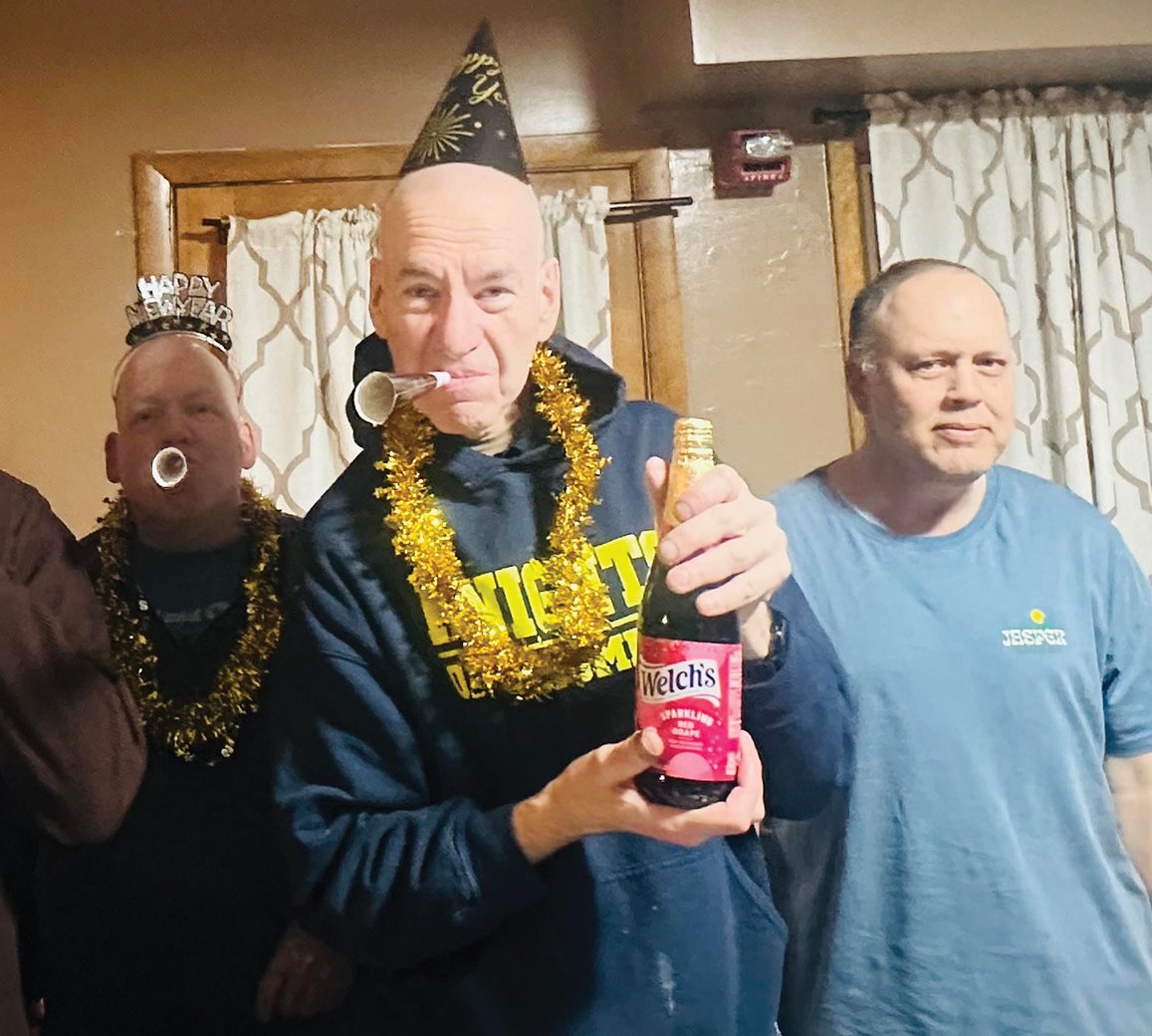 Group of men celebrating New Year's Eve with party hats, tinsel, and noise makers in a cozy room, one holding a bottle of Welch's sparkling drink.