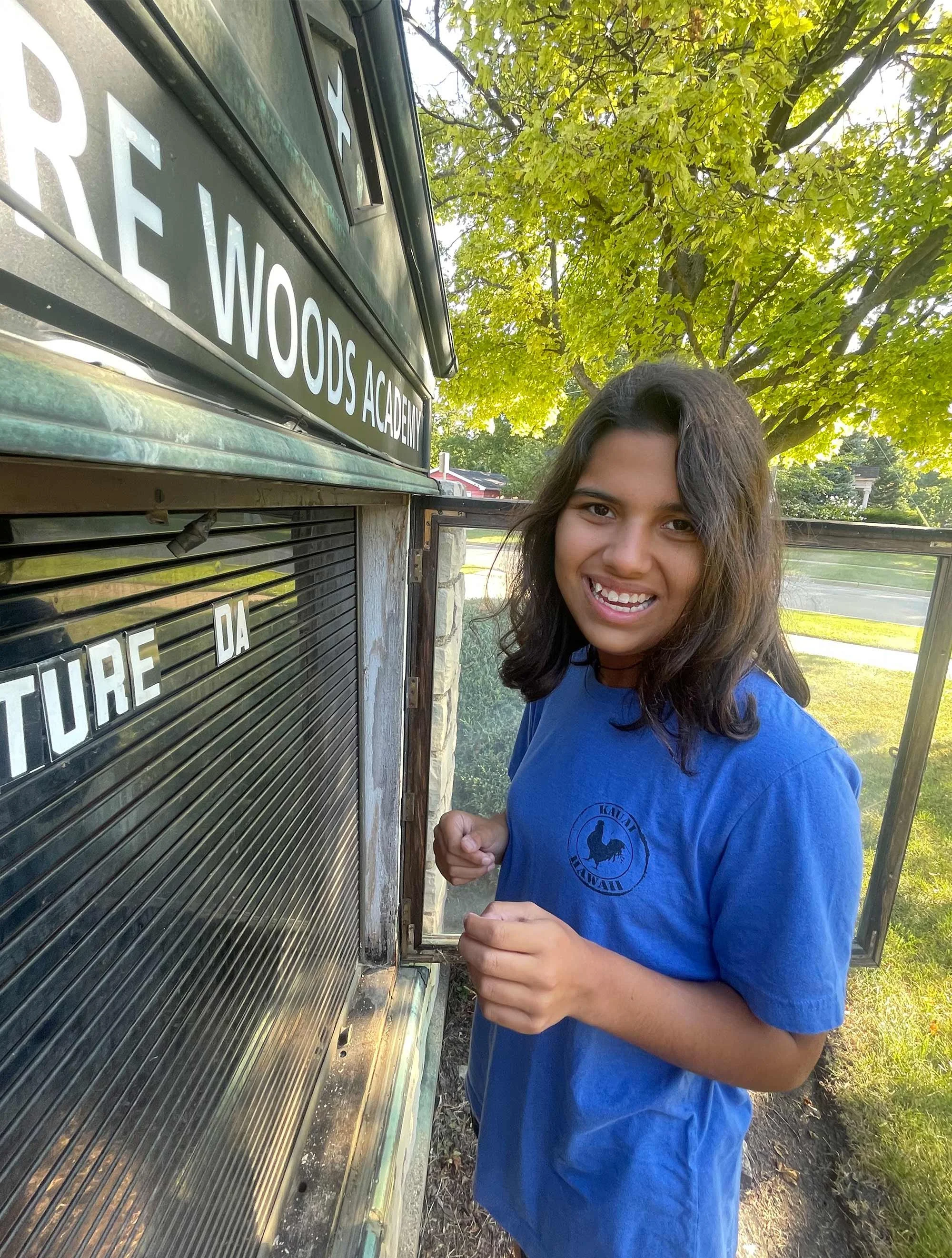 A young woman with long dark hair, smiling, wearing a blue T-shirt, standing outside in front of a letterboard sign that says "CLAR WOODS ACADEMY." She is holding letters to place on the board.