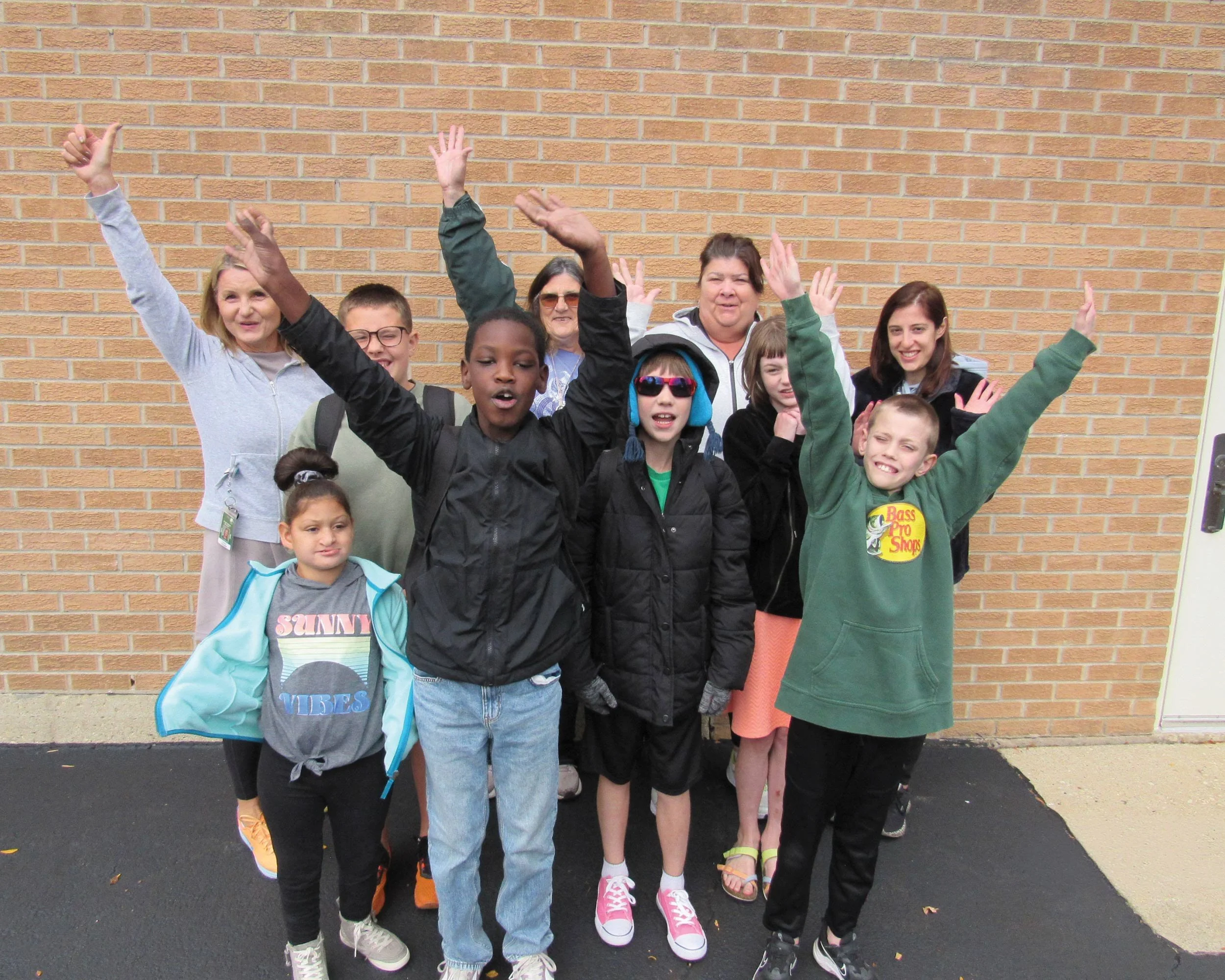 Group of children and women standing outdoors in front of a brick wall, celebrating with raised hands and smiling.
