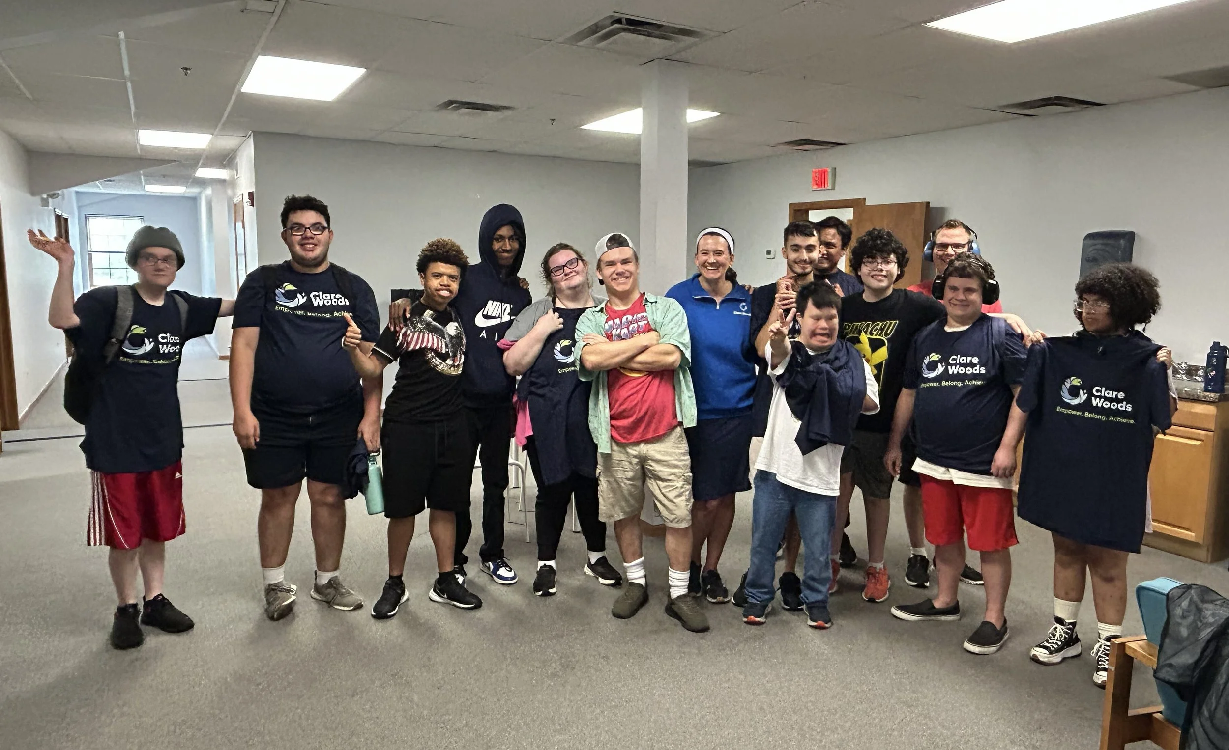 Group of people smiling and standing together indoors, some with headphones, wearing Clare Woods T-shirts.