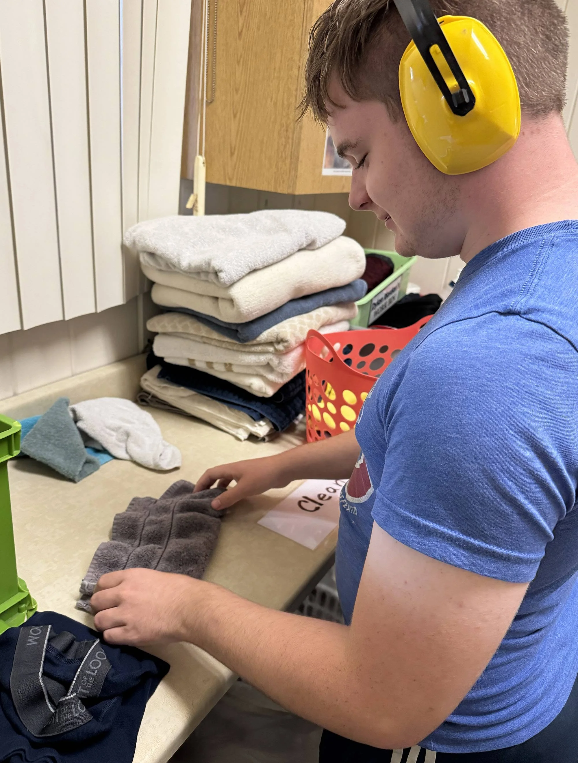 Young man folding clothes at a table, wearing yellow safety earmuffs and a blue T-shirt, with a pile of folded towels and laundry baskets in the background.