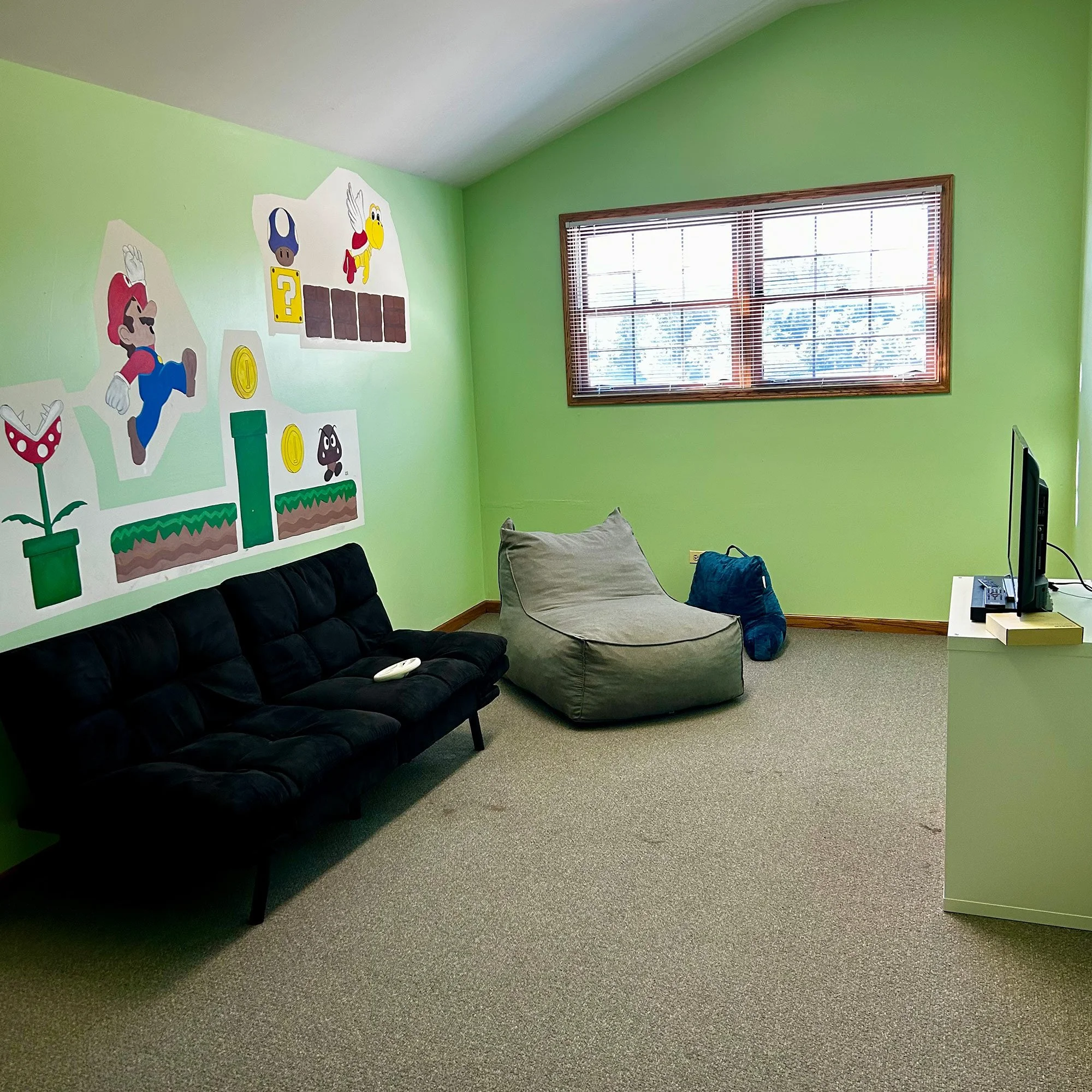 An image of the Day Program room. Room with green walls, wall decoration featuring Super Mario characters and elements, window with blinds, black couch, beige armchair, and a carpeted floor.