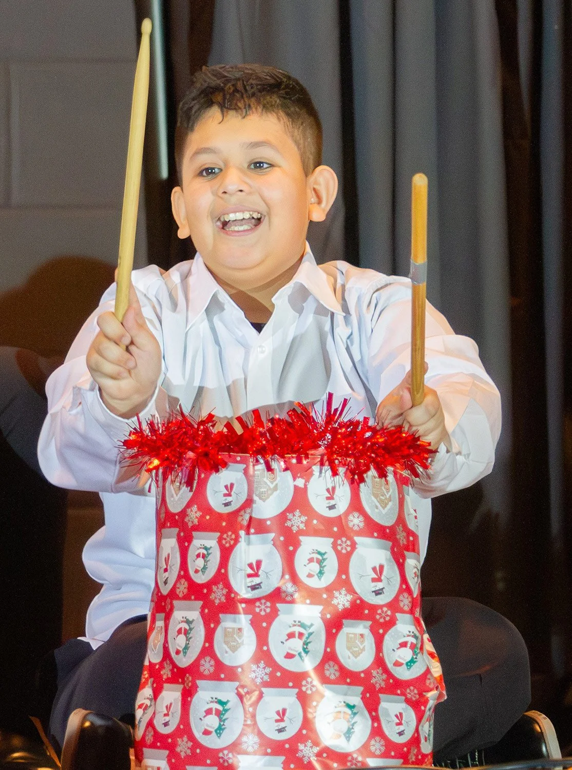 A young smiling boy in a white shirt, holding drumsticks and playing on a drum decorated with snowmen and snowflakes.
