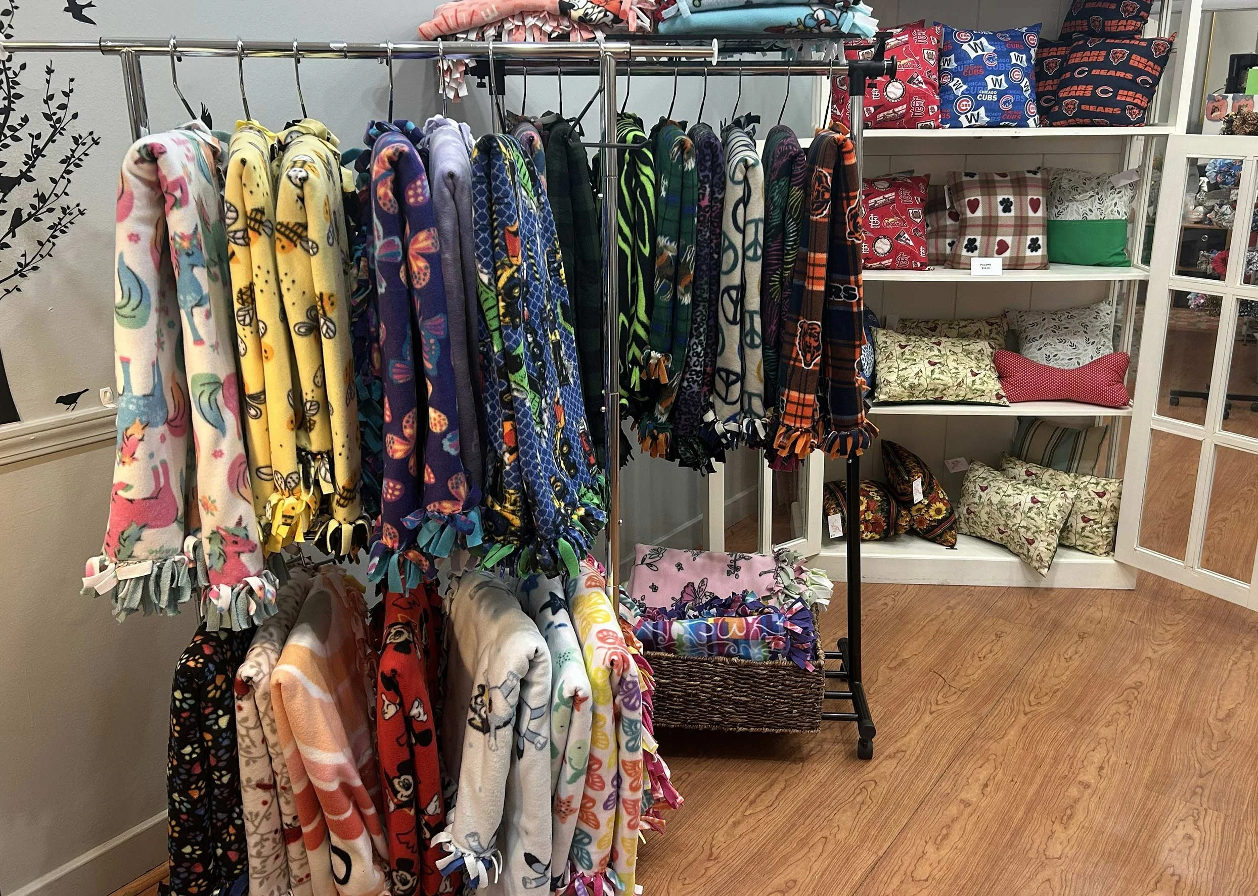 Display of colorful fleece blankets with various patterns hanging on a clothing rack. Shelves with more blankets and pillows are visible in the background.