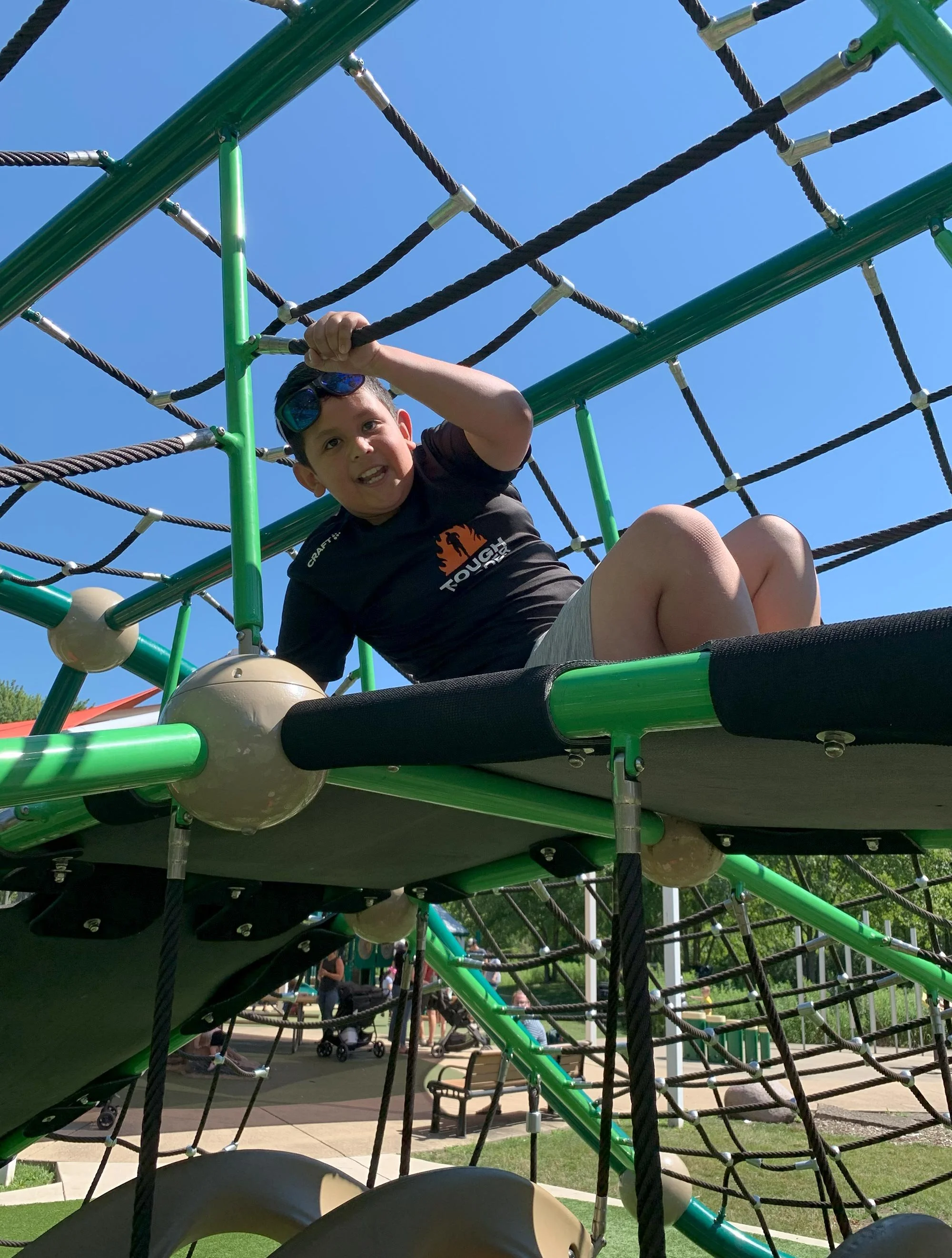 A young boy wearing sunglasses and a black t-shirt is climbing on a green and black playground structure made of ropes and metal bars, with a blue sky in the background.