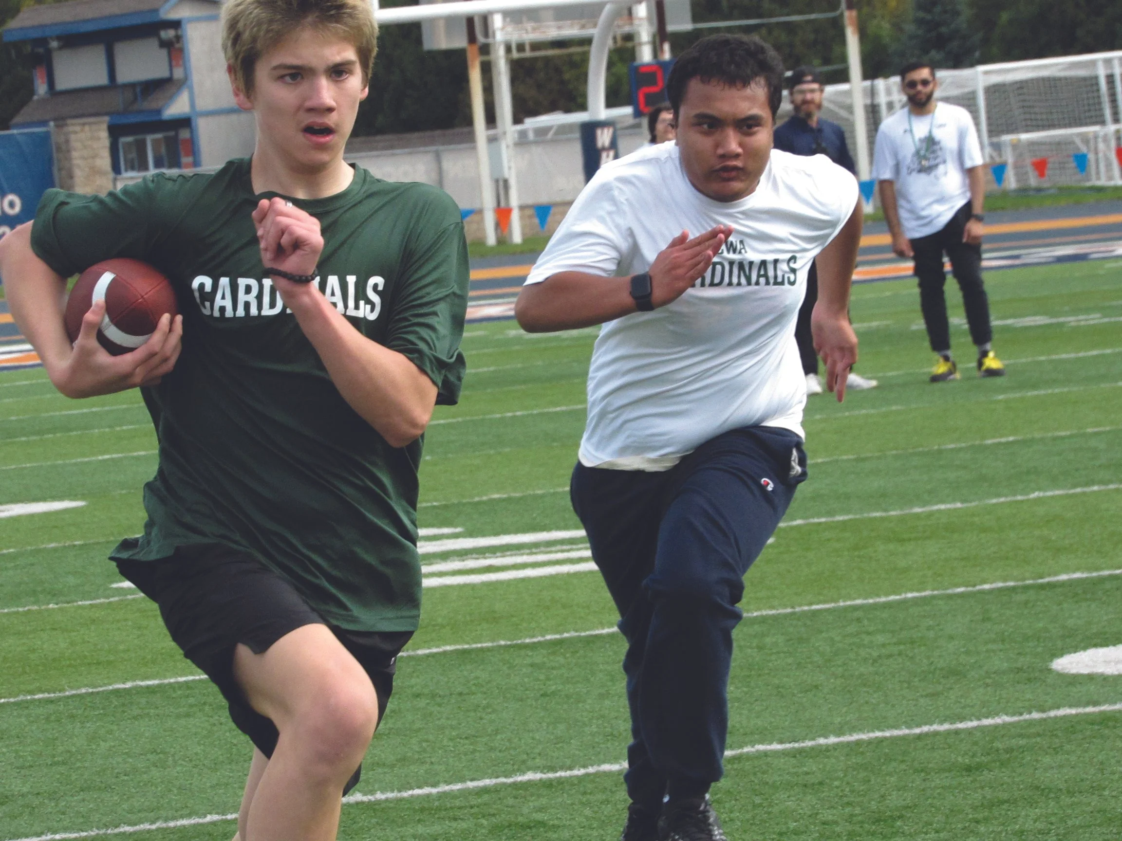 Two young male athletes running on a track, one holding a football, wearing sports shirts with the word 'Cardinals,' on a sports field with goalposts and a scoreboard in the background.