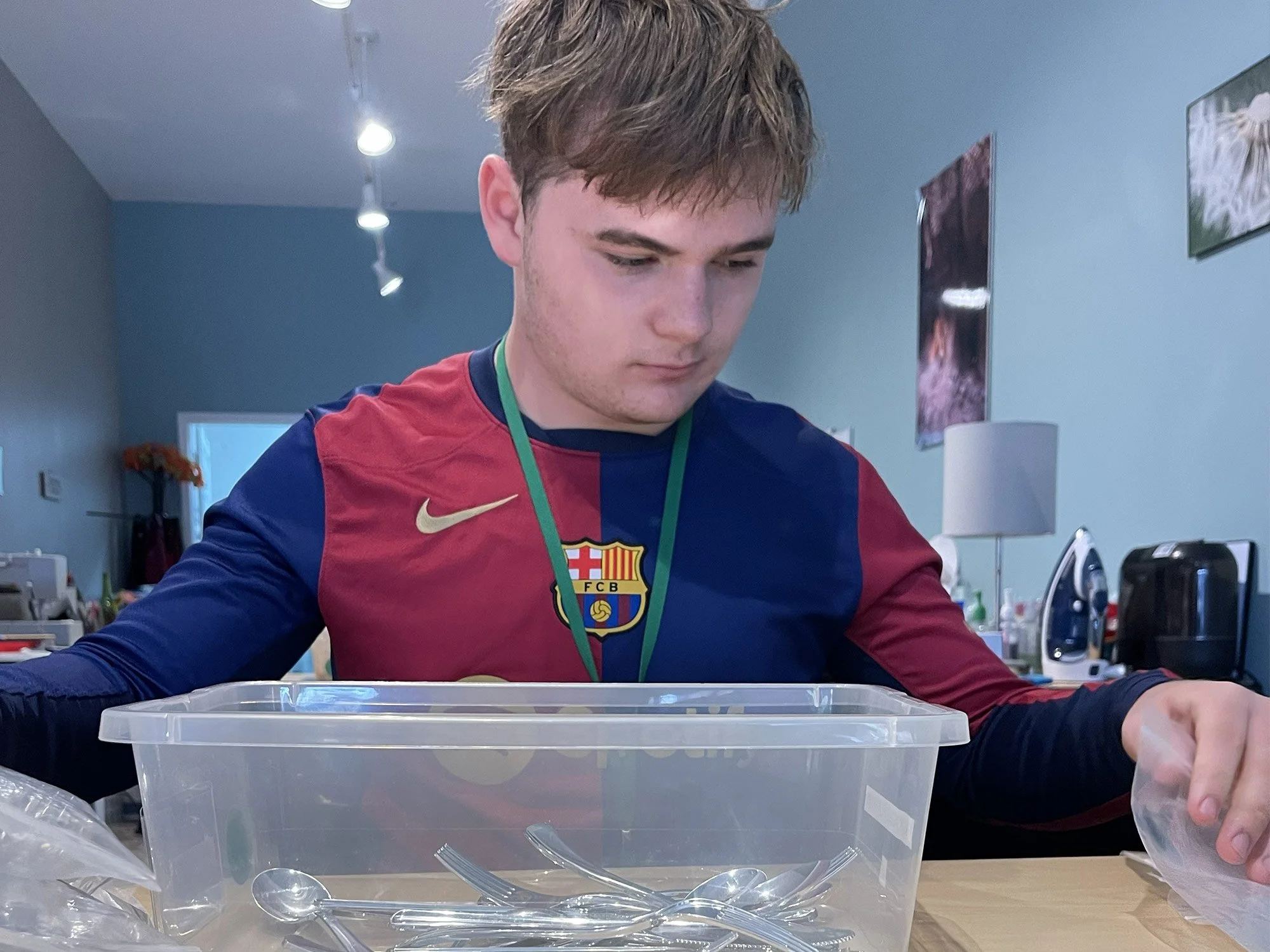 A young man with light brown hair wearing a red and blue sports jersey, sitting at a table with plastic containers filled with small objects, inside a room with artwork on the walls and bright ceiling lights.