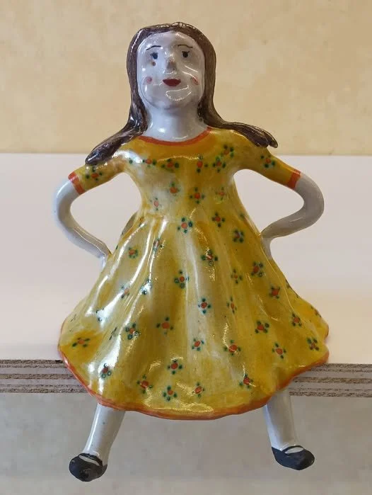 A ceramic figurine of a girl in a yellow dress with floral patterns, standing with her hands on her hips.