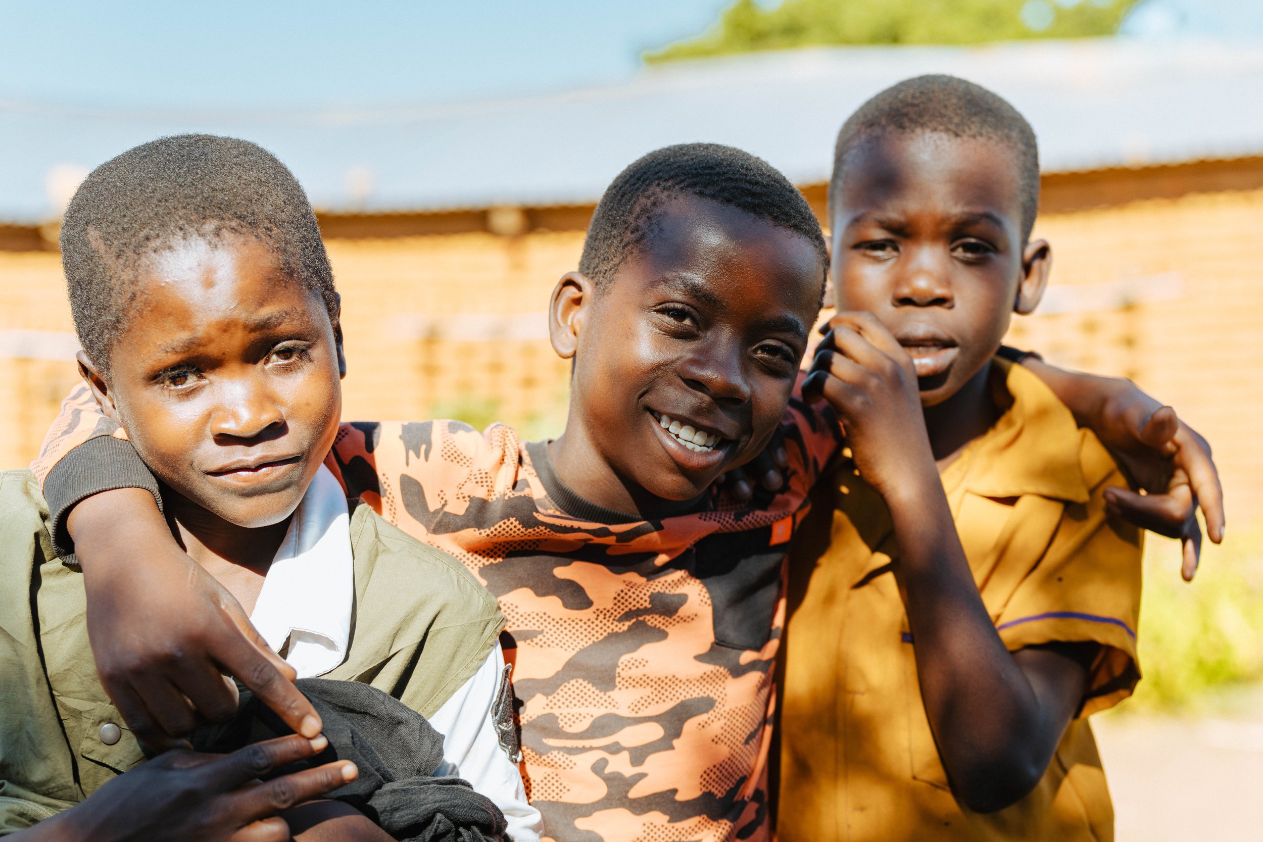 Three boys with short hair smiling and sitting close together outdoors.