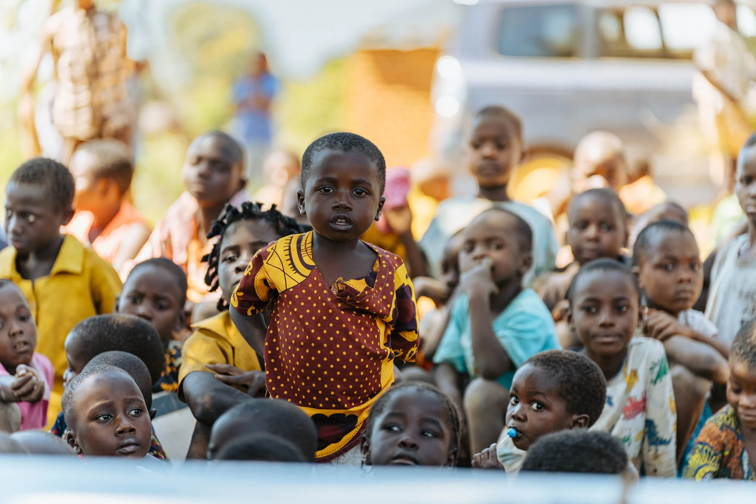 A young girl with short hair dressed in a colorful dress stands among a crowd of children, most of whom are sitting and looking in different directions, outdoors with a blurred vehicle and people in the background.