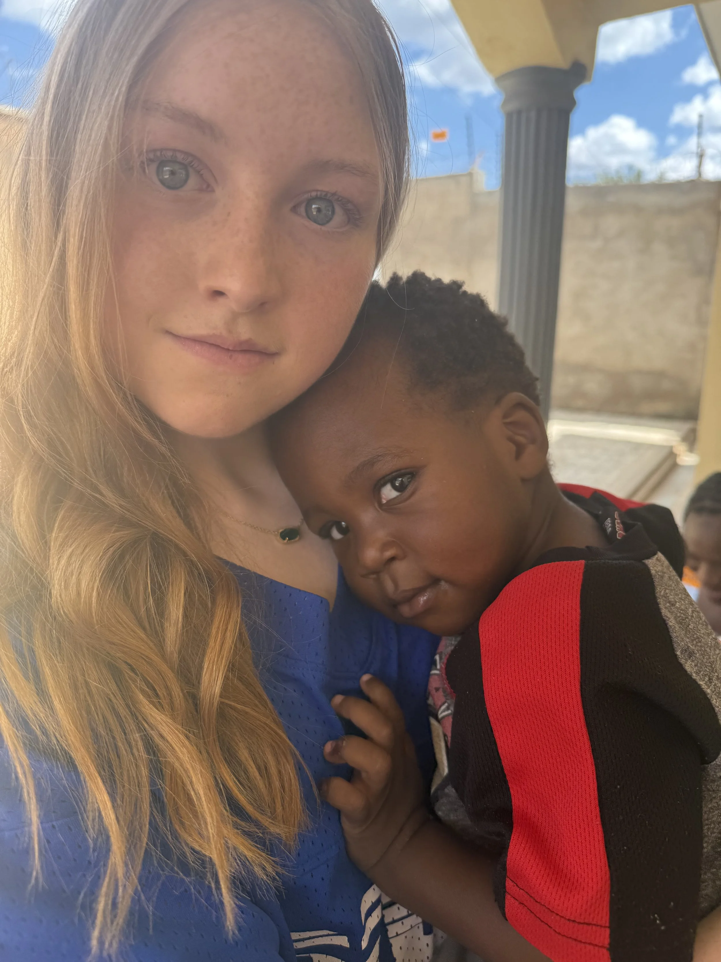 A young woman with long blonde hair and freckles takes a selfie with a young boy who has dark hair and a dark complexion, both are smiling slightly and are outdoors with a bright blue sky and columns in the background.