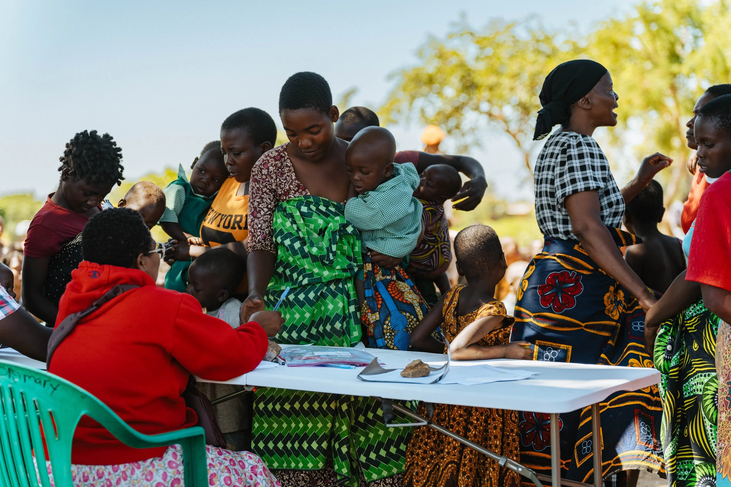 Women and children gathered around a table outdoors participating in an event, with some women holding children and a person seated at the table writing or signing documents.