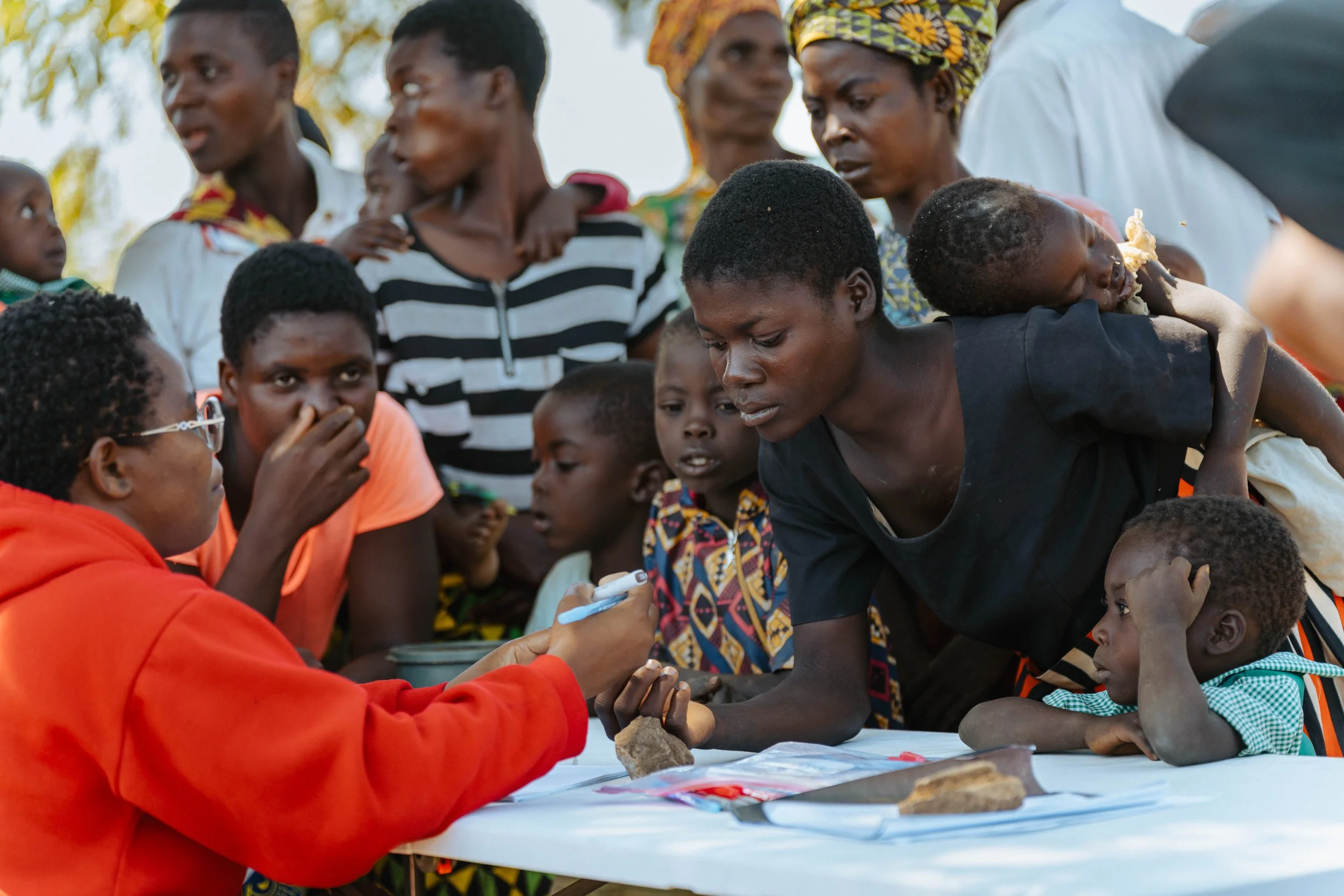 A group of African women and children gather around a table outdoors, engaging with a woman in a red jacket who is signing a document or filling out a form. The scene appears to be a community or outreach event under natural daylight.