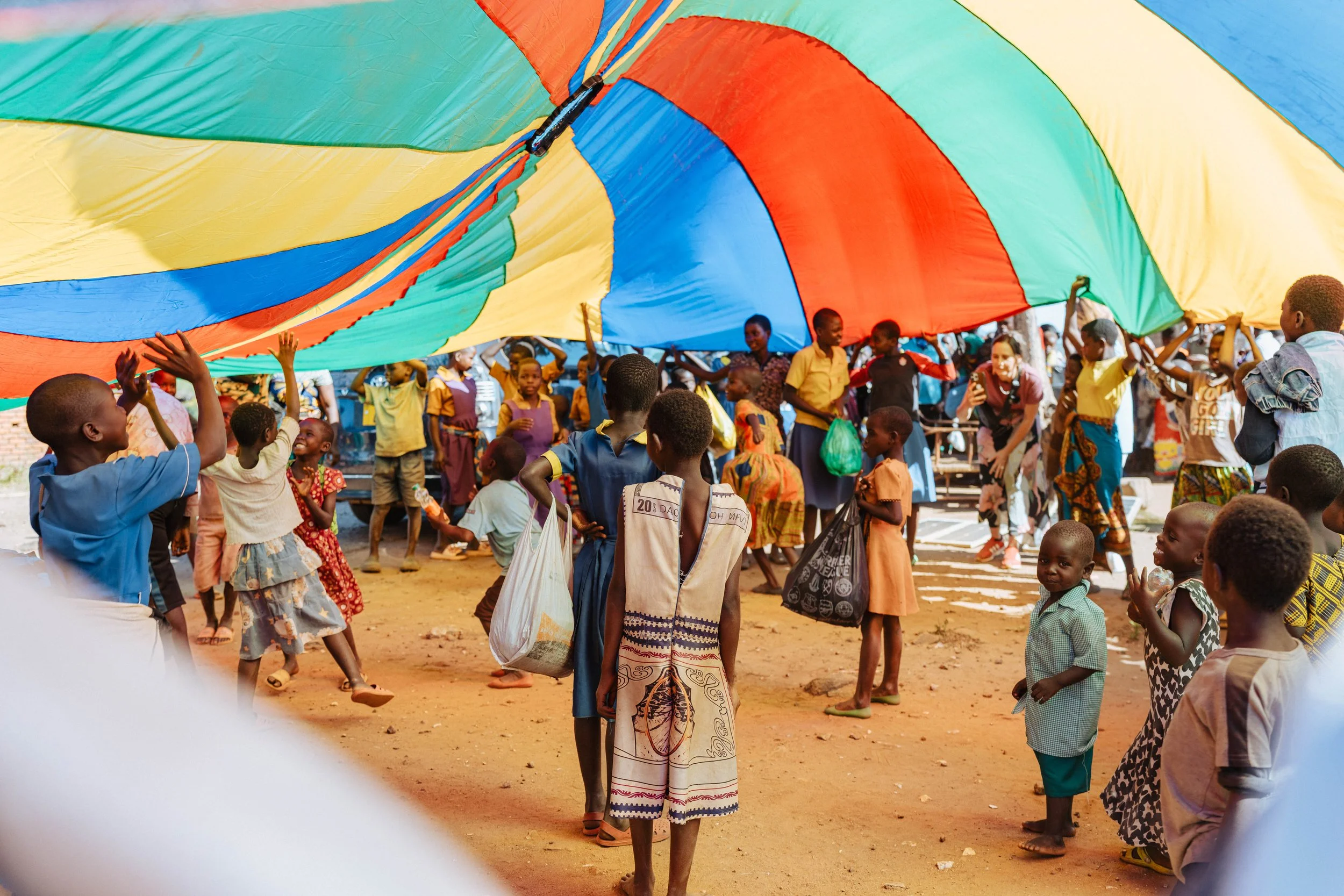 Children playing with a large colorful parachute outdoors, with some children holding the edges and others underneath, smiling and having fun.