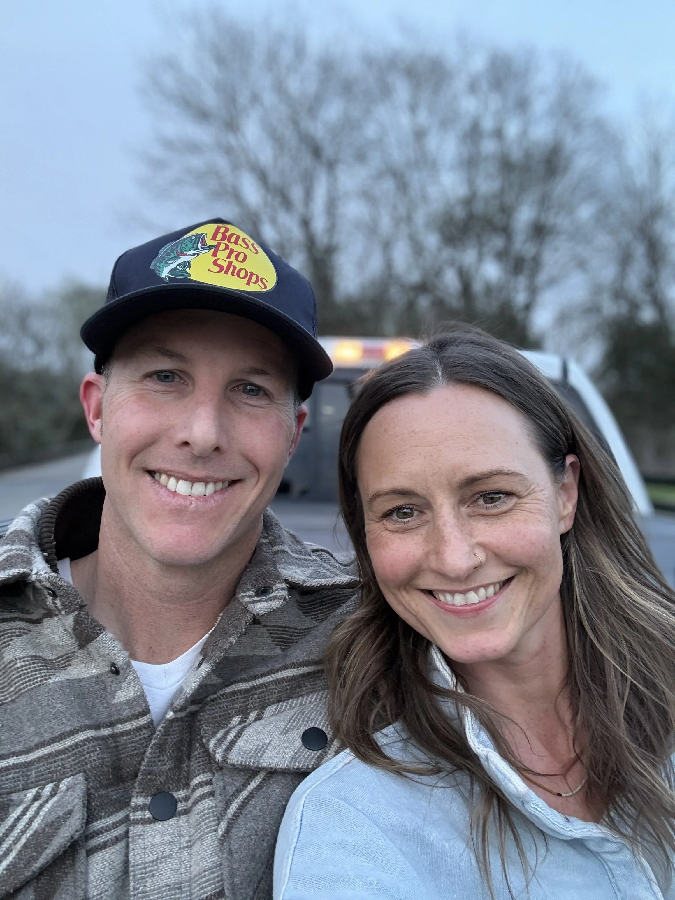 A cheerful man and woman are taking a selfie outdoors, smiling at the camera. The man is wearing a Bass Pro Shops hat and a plaid jacket, and the woman has long brown hair and a nose ring. A vehicle and a tree are visible in the background.