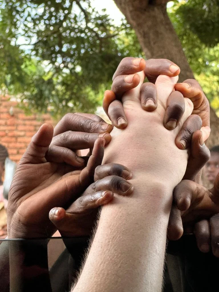 Multiple dark-skinned hands grasp and hold a single light-skinned hand in a group gesture. The scene is outdoors with green trees and a brick wall in the background.
