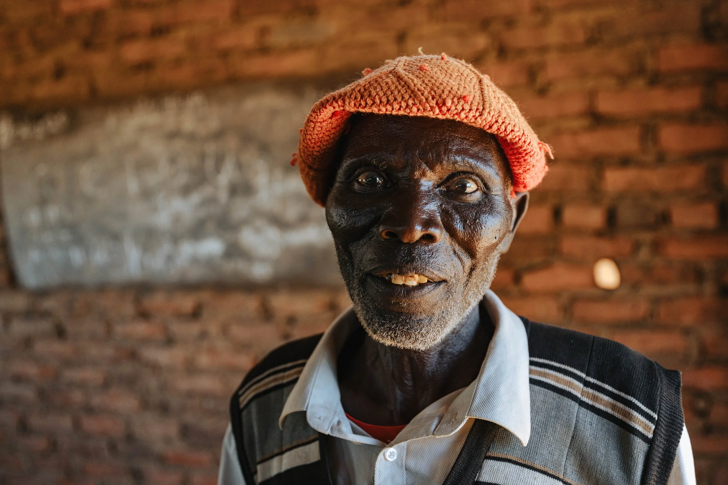 An elderly man with dark skin wearing a pink knitted hat, a white collared shirt beneath a dark sweater with striped details, standing in front of a brick wall.
