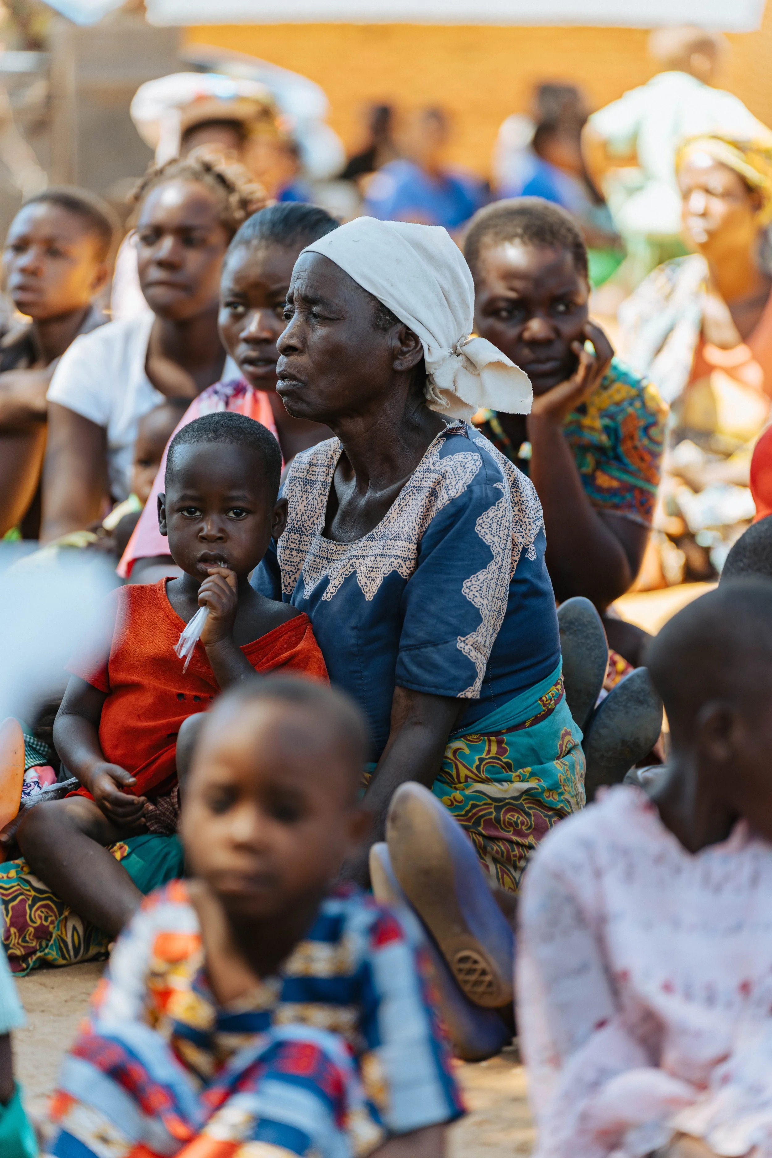 A group of African children and women sitting on the ground outdoors, listening attentively to an event or speaker. Some are wearing colorful clothing and traditional head wraps, while others are in casual attire.