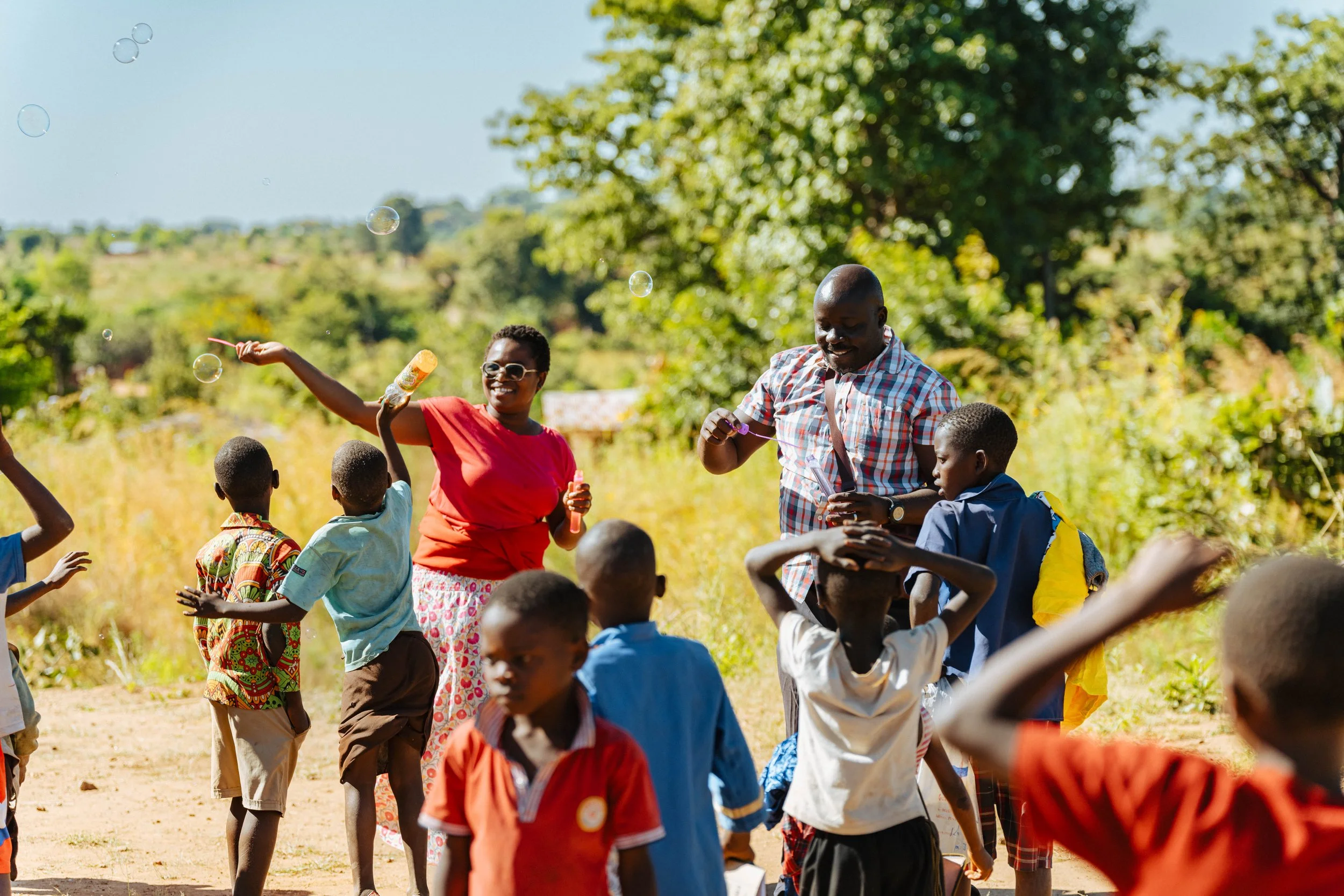 Children and adults playing outdoors in a sunny field with trees in the background, some holding bubbles and bubbles floating in the air, enjoying a lively gathering.