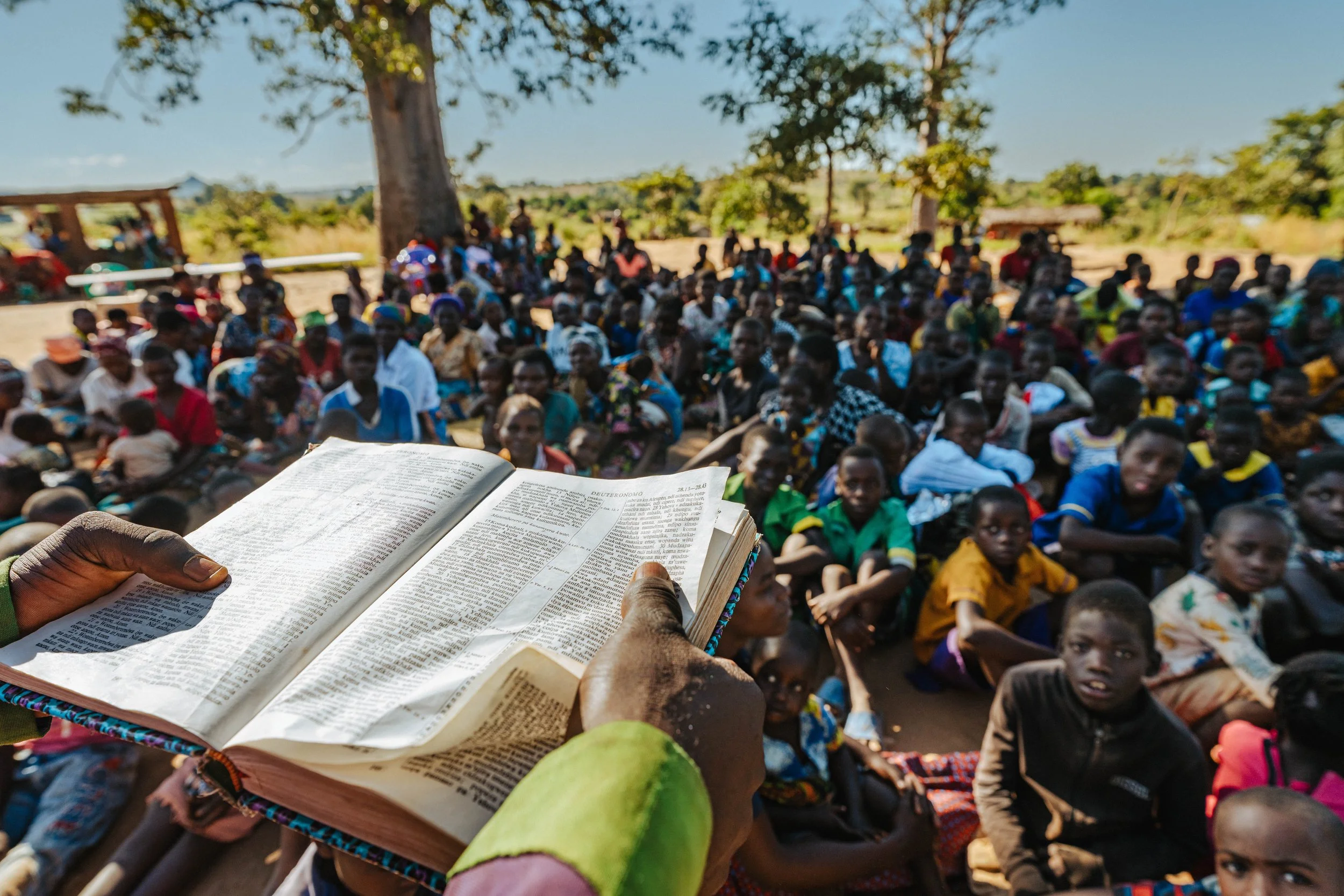 A person holding an open Bible and reading to a large crowd of people sitting outdoors under trees, with a rural landscape in the background.