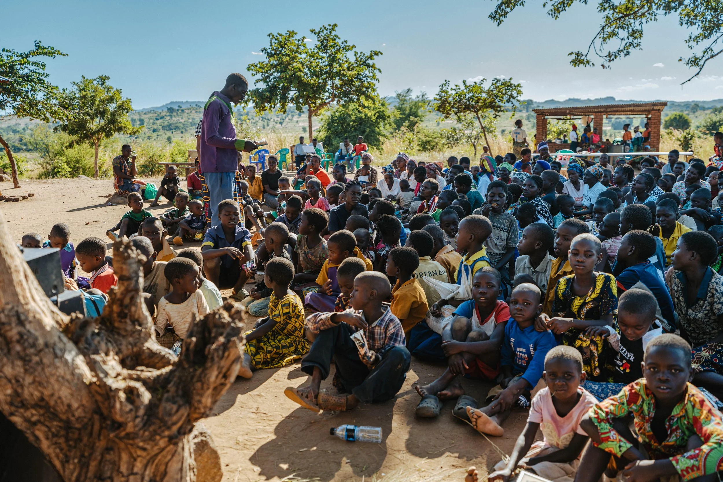A large gathering of children and a few adults outdoors, some sitting on the ground and others standing, under trees with a brick shelter in the background, in a rural setting with hills and greenery.
