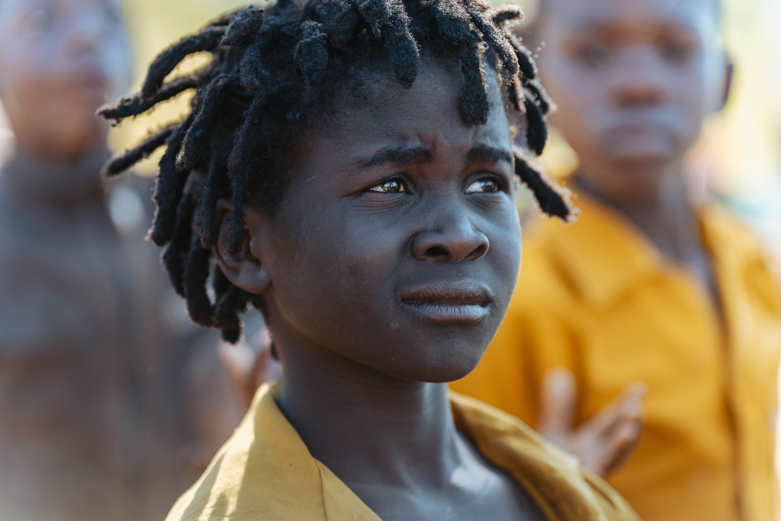 Close-up of a young person with dark skin and dreadlocks, wearing a yellow shirt, with a thoughtful expression, and two other blurred individuals in the background.