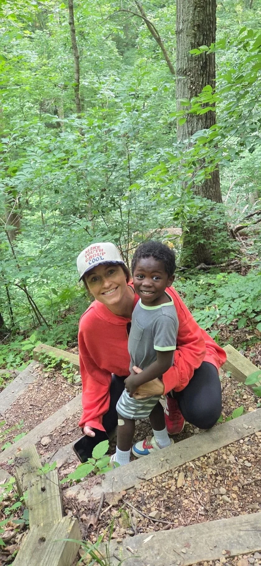A smiling woman in a gray cap and red jacket is kneeling on a trail in the woods, holding a young boy who is standing in front of her, wearing a gray t-shirt, striped shorts, and sneakers, all surrounded by lush green forest.