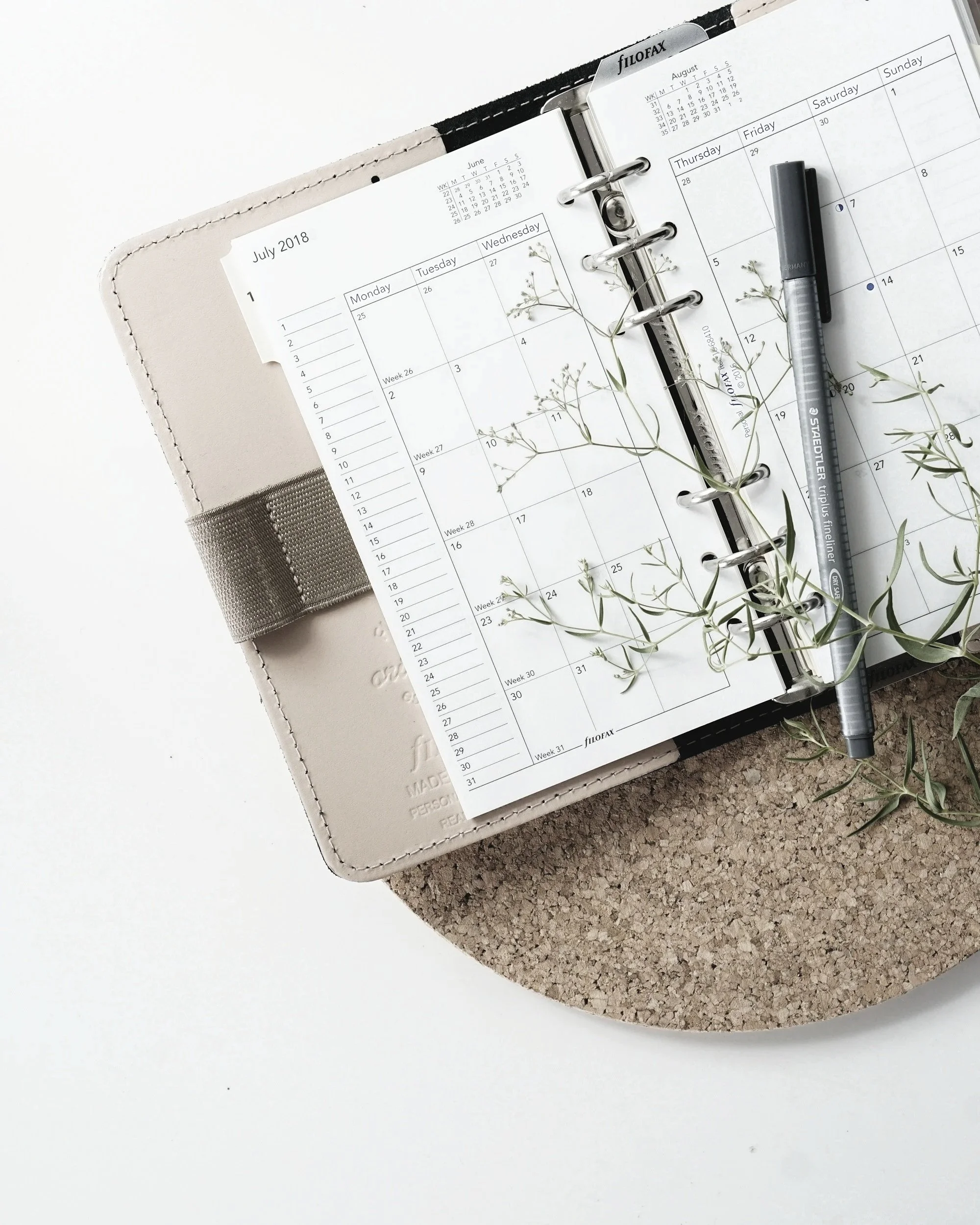 Open planner displaying calendar pages, secured with metal rings, on top of a beige bag with stitched edges, adorned with sprigs of small white flowers, set on a surface with white and textured brown sections, with a gray marker placed on the planner.