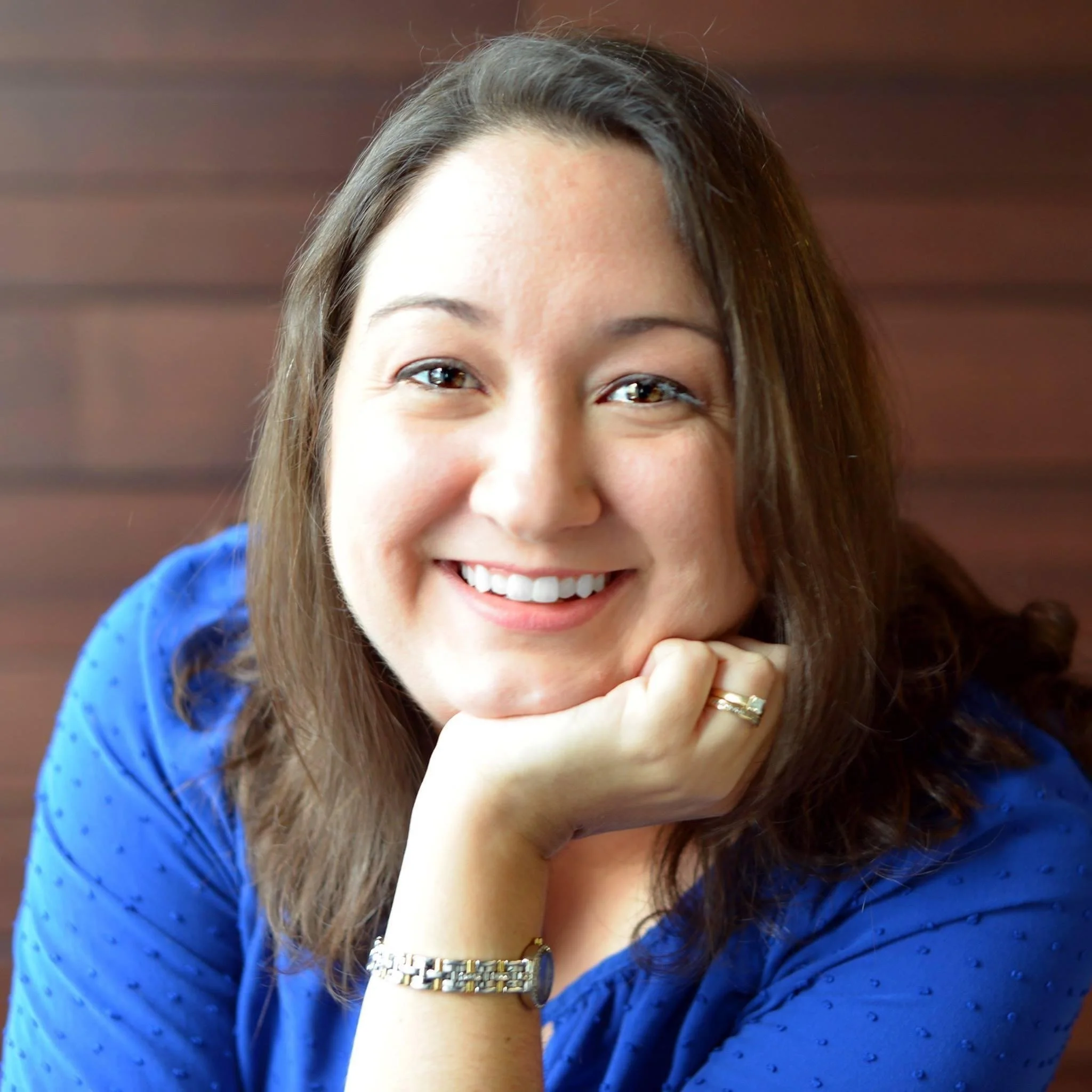 Close-up of Heather Ackmann, smiling woman with brown hair, wearing a blue shirt, a gold bracelet, a ring, and resting her chin on her hand.
