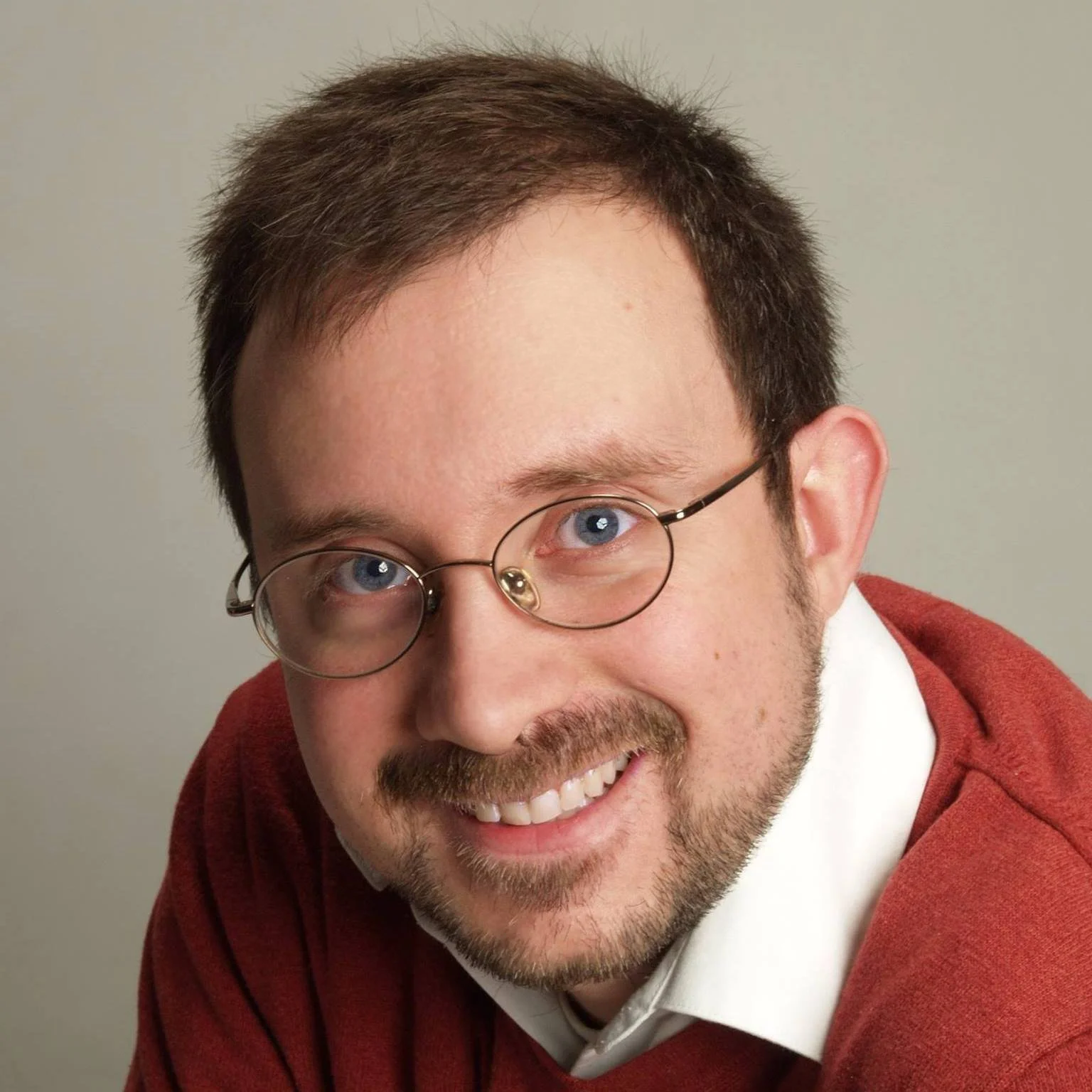 Close-up of Alan Ackmann, a smiling man with glasses, brown hair, and a beard, wearing a red sweater over a white shirt.