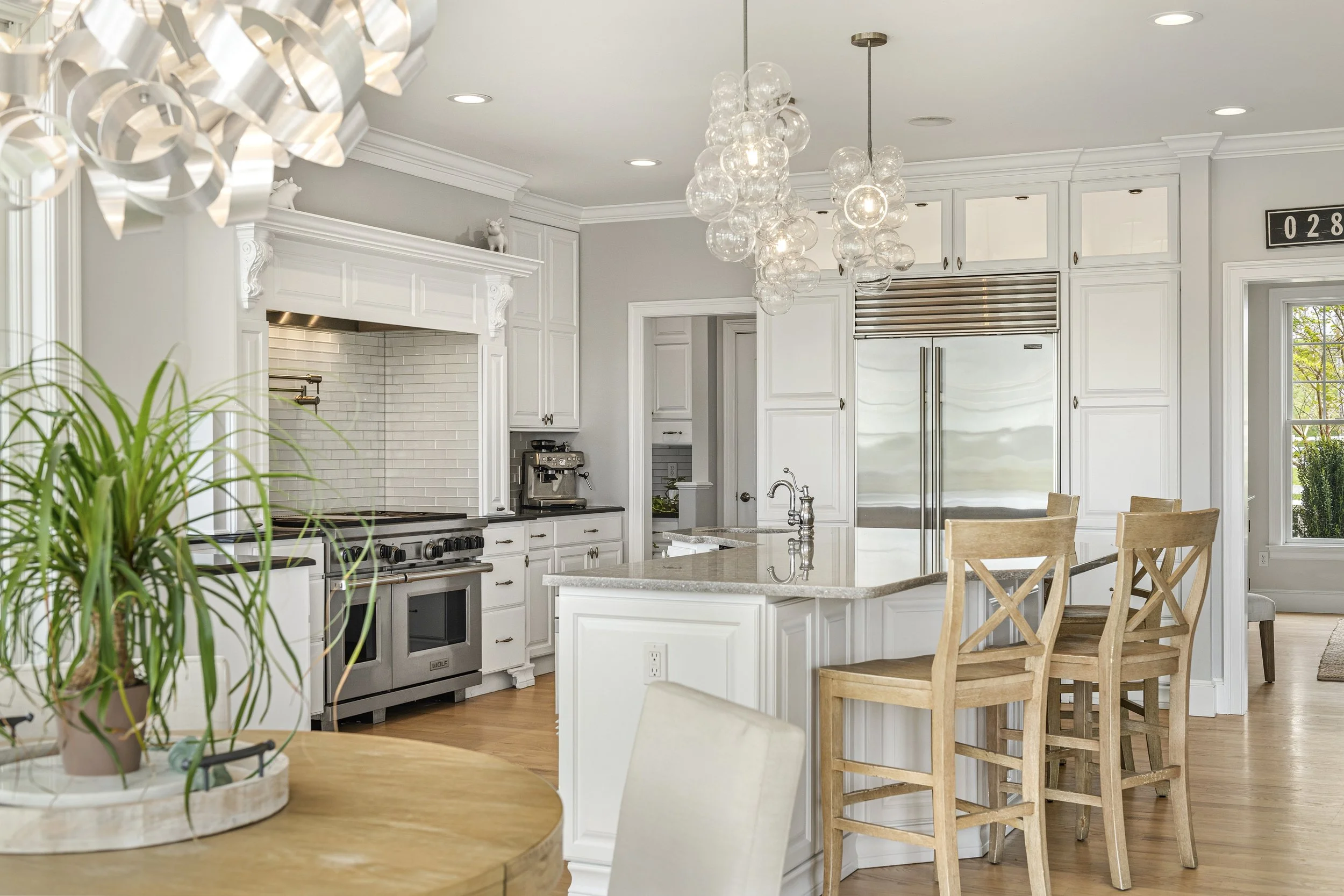 Modern kitchen with white cabinetry, stainless steel appliances, a kitchen island, and wooden chairs. Interior decor includes glass chandeliers and decorative items on top of the cabinets.