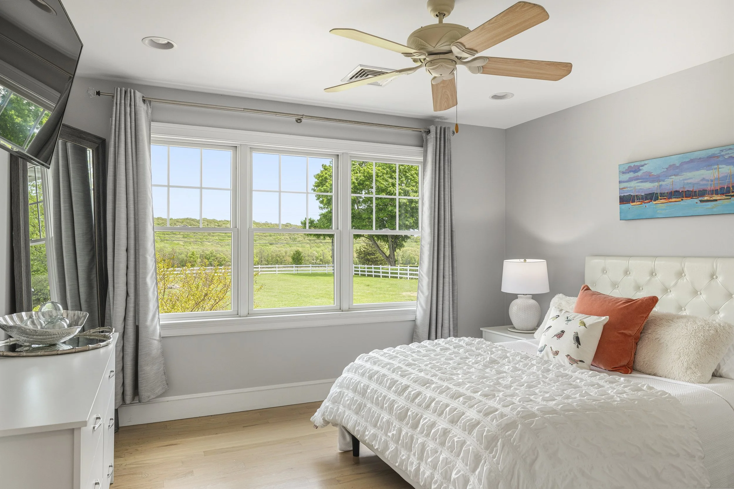 A bedroom with large windows showing a green landscape outside, white curtains, a ceiling fan, a white bed with decorative pillows, a white nightstand with a lamp, and a colorful painting of boats on the wall.