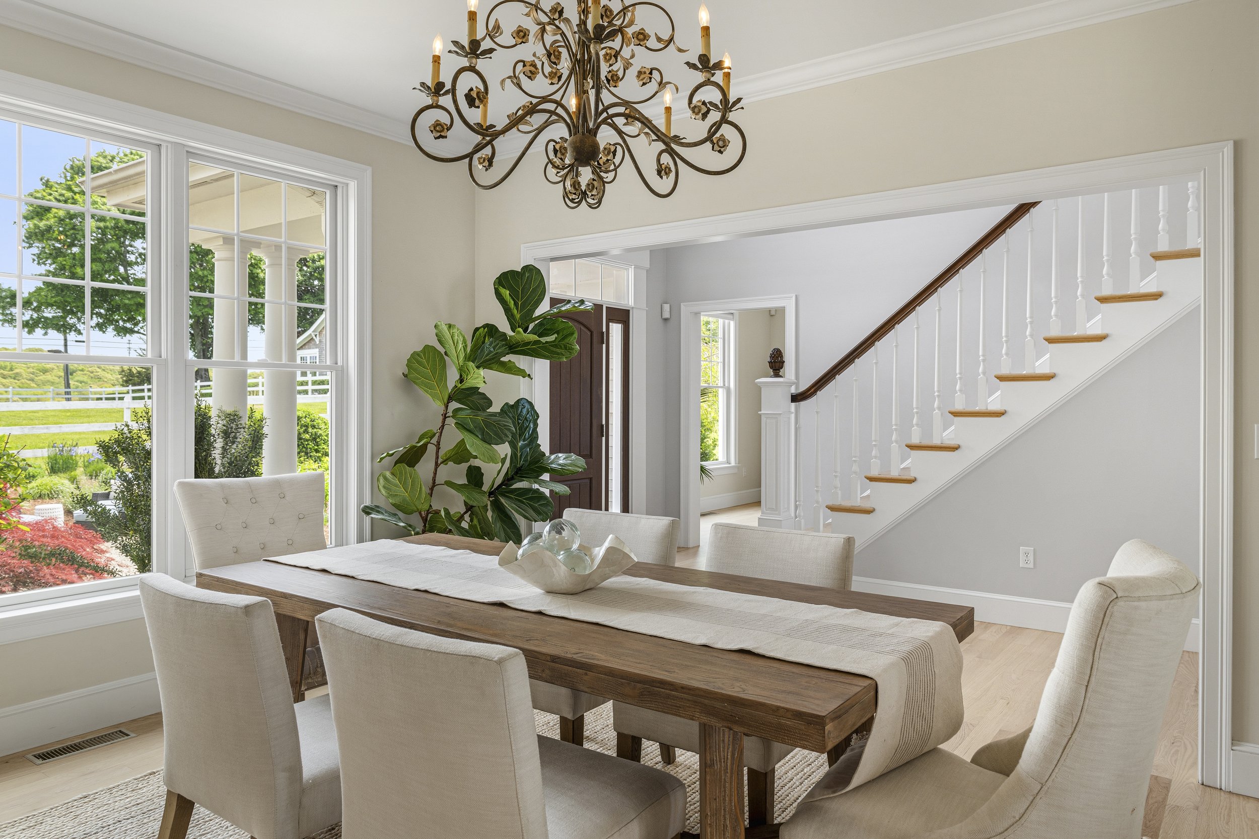 A dining room with large windows, a wooden dining table with a beige table runner, surrounded by six upholstered chairs. There is a large green plant near the windows and a chandelier hanging from the ceiling. The room opens to a staircase and a fron