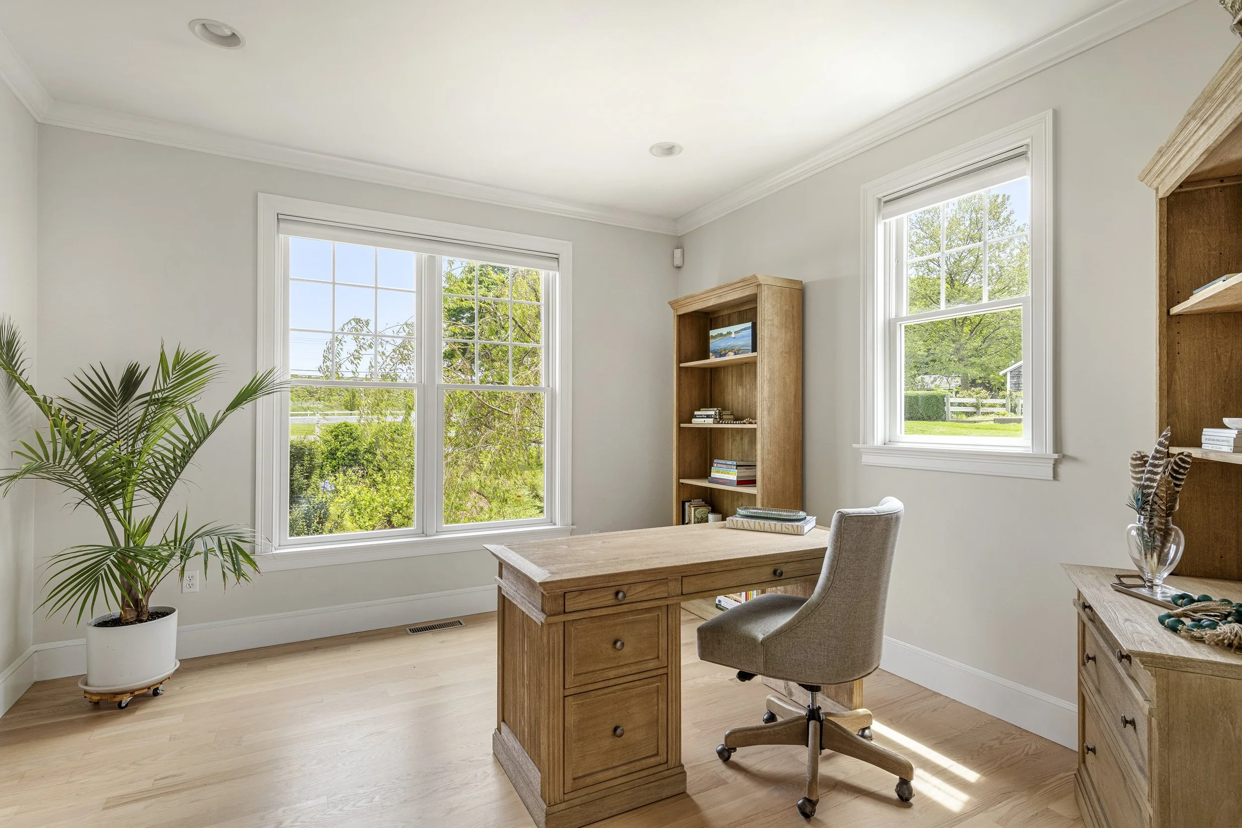 A bright home office with large windows showing green trees outside, wooden furniture including a desk with drawers, a tall bookshelf with books and decor, a gray office chair, a potted plant, and a decorative feather in a glass vase.