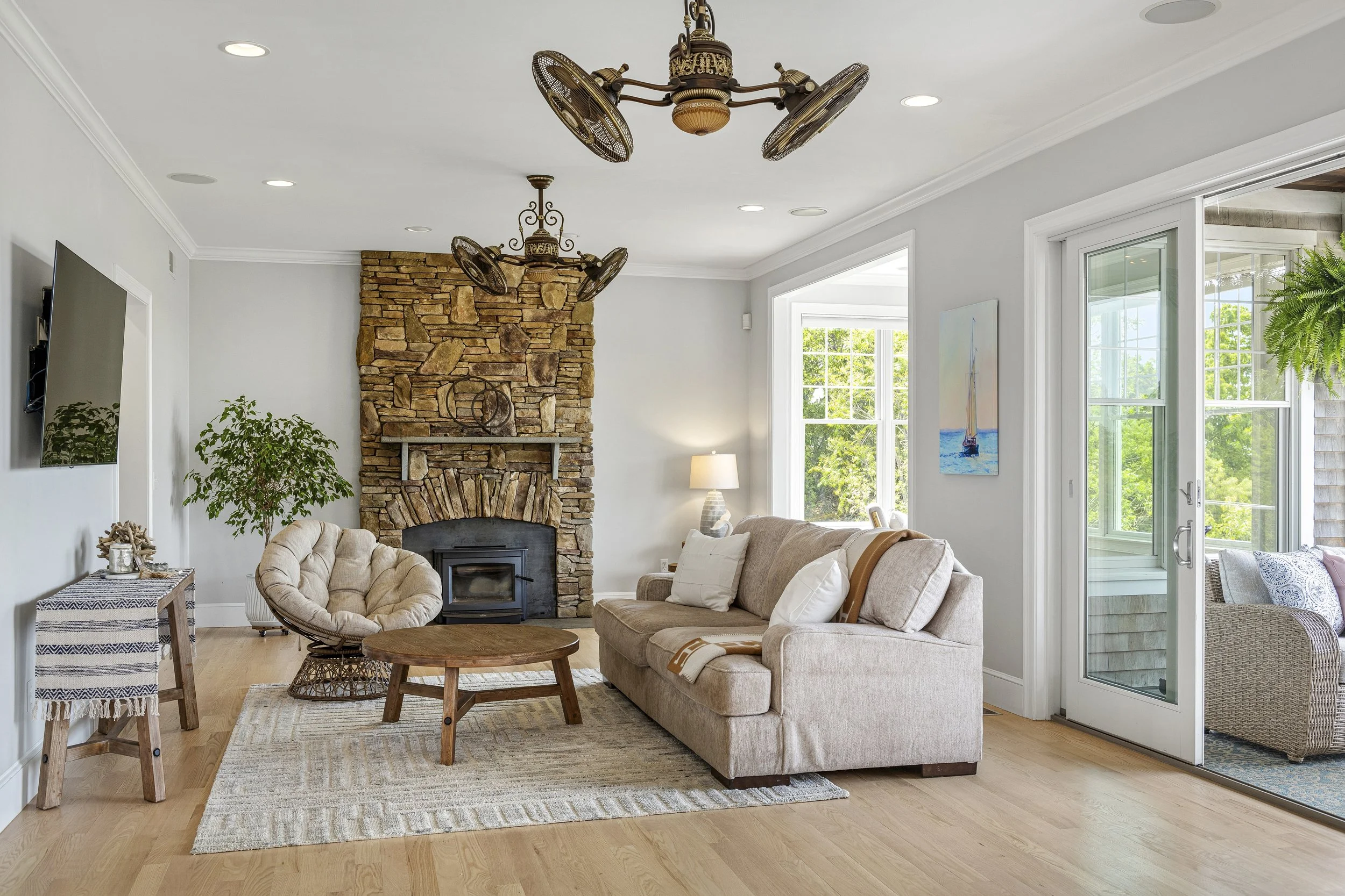 Bright living room with a stone fireplace, beige sofa, cozy armchair, wooden coffee table, large windows with lush greenery outside, ceiling fans, and decorative artwork.