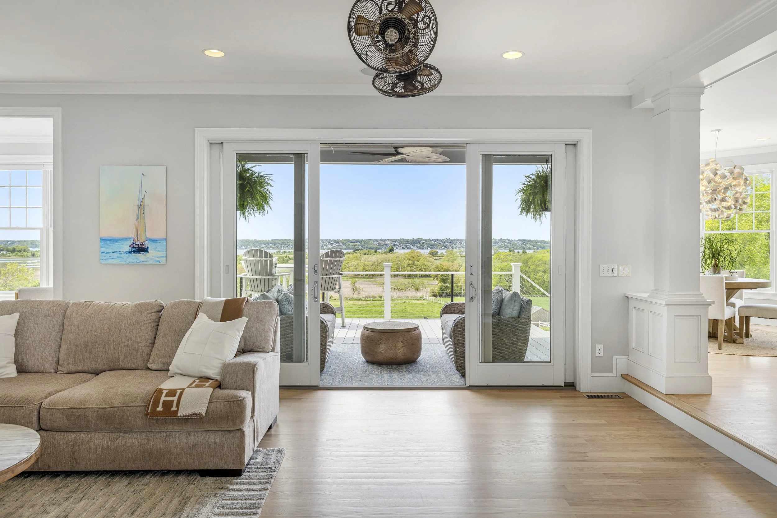 Living room with beige sofa, white pillows, and a coffee table on a woven rug, opening to a deck with outdoor chairs and a view of trees and water.