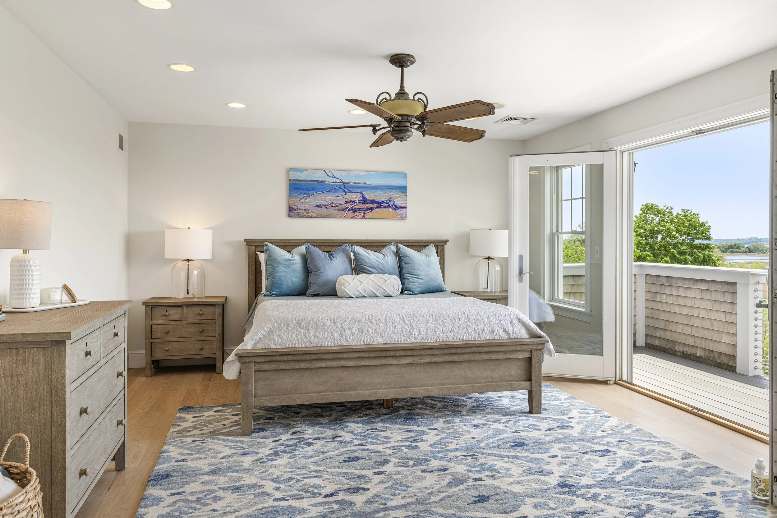 Bedroom with a wooden bed, blue pillows, a painting of a beach with driftwood, two nightstands with lamps, an open balcony door showing greenery outside, and a ceiling fan.