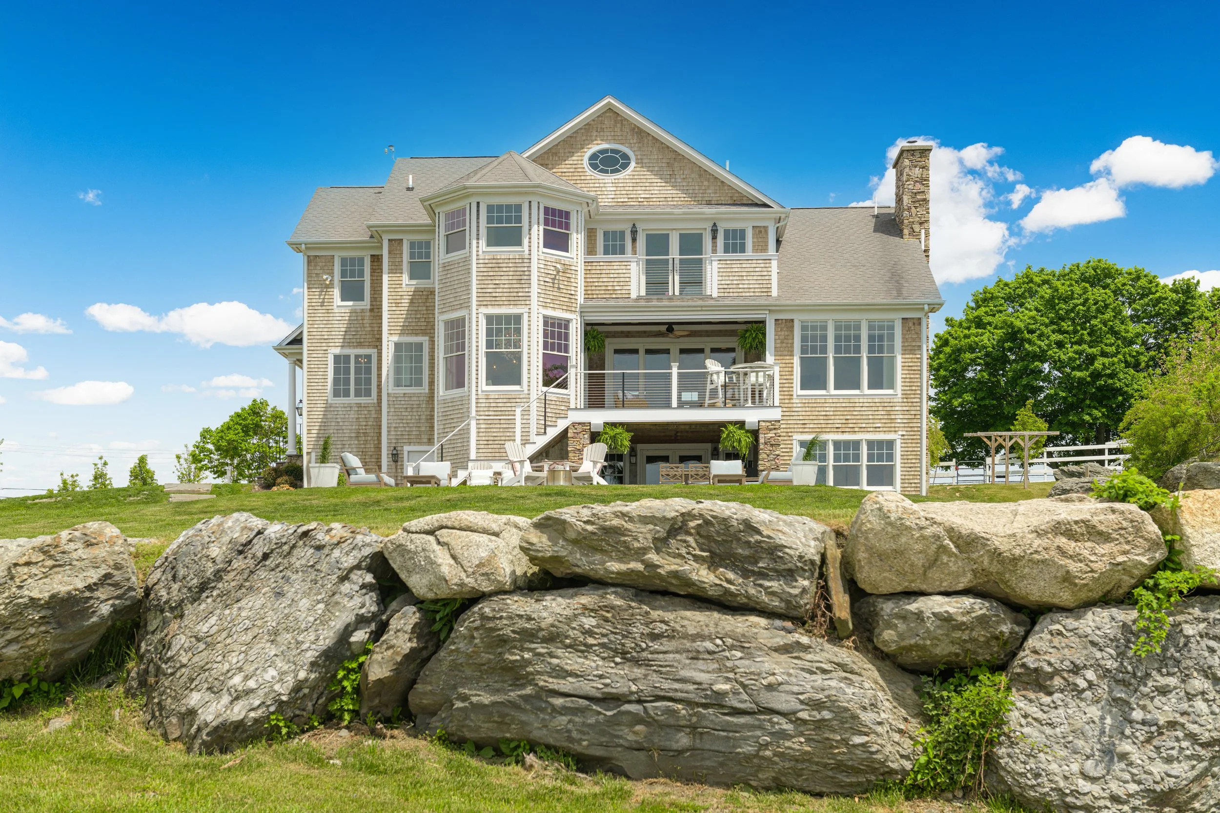 A spacious house with multiple stories, large windows, and a balcony, situated behind a low stone wall with a lush green lawn and trees, under a bright blue sky with some clouds.