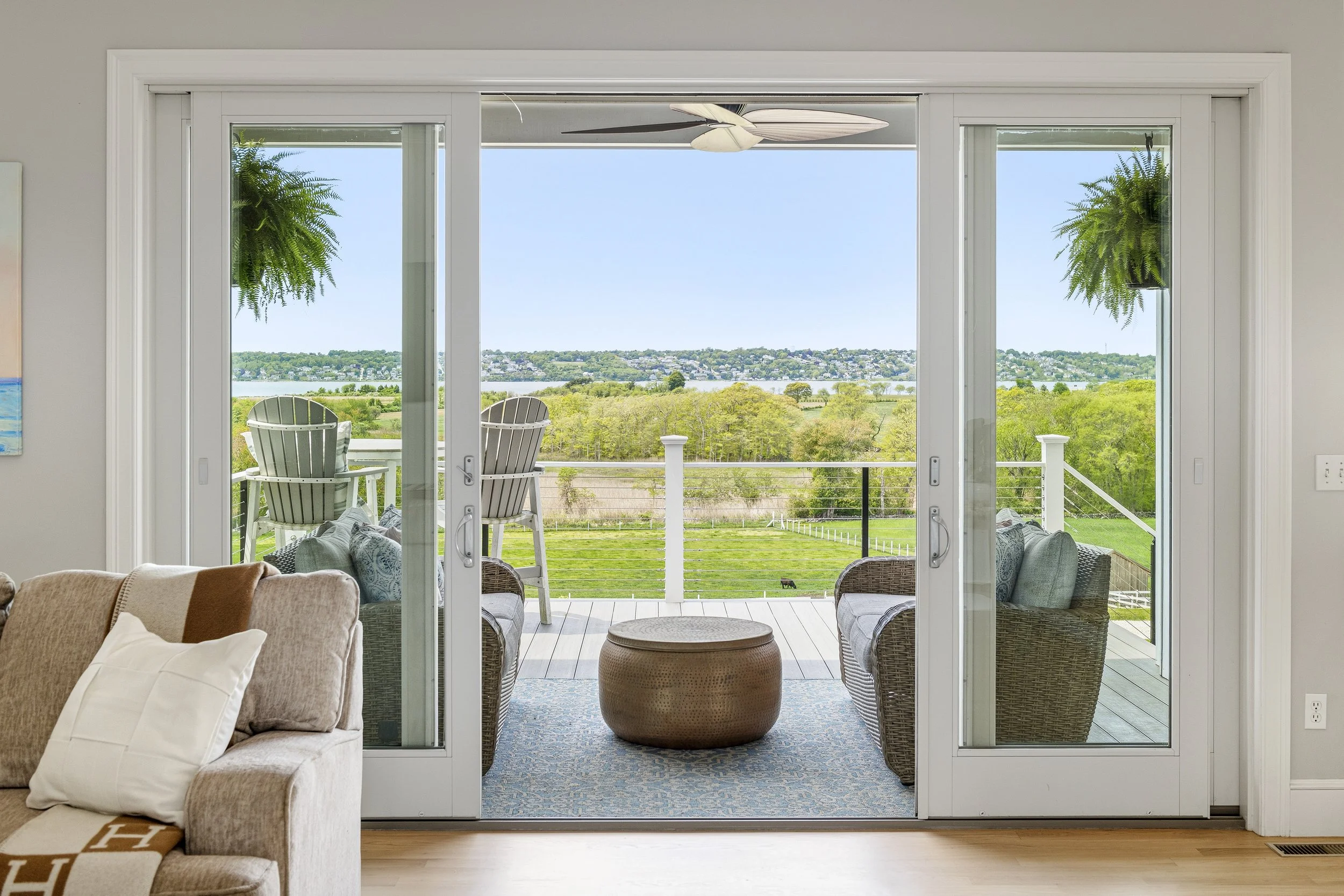 Living room with sliding glass door leading to a balcony overlooking a green landscape and water, with outdoor chairs and hanging potted plants.