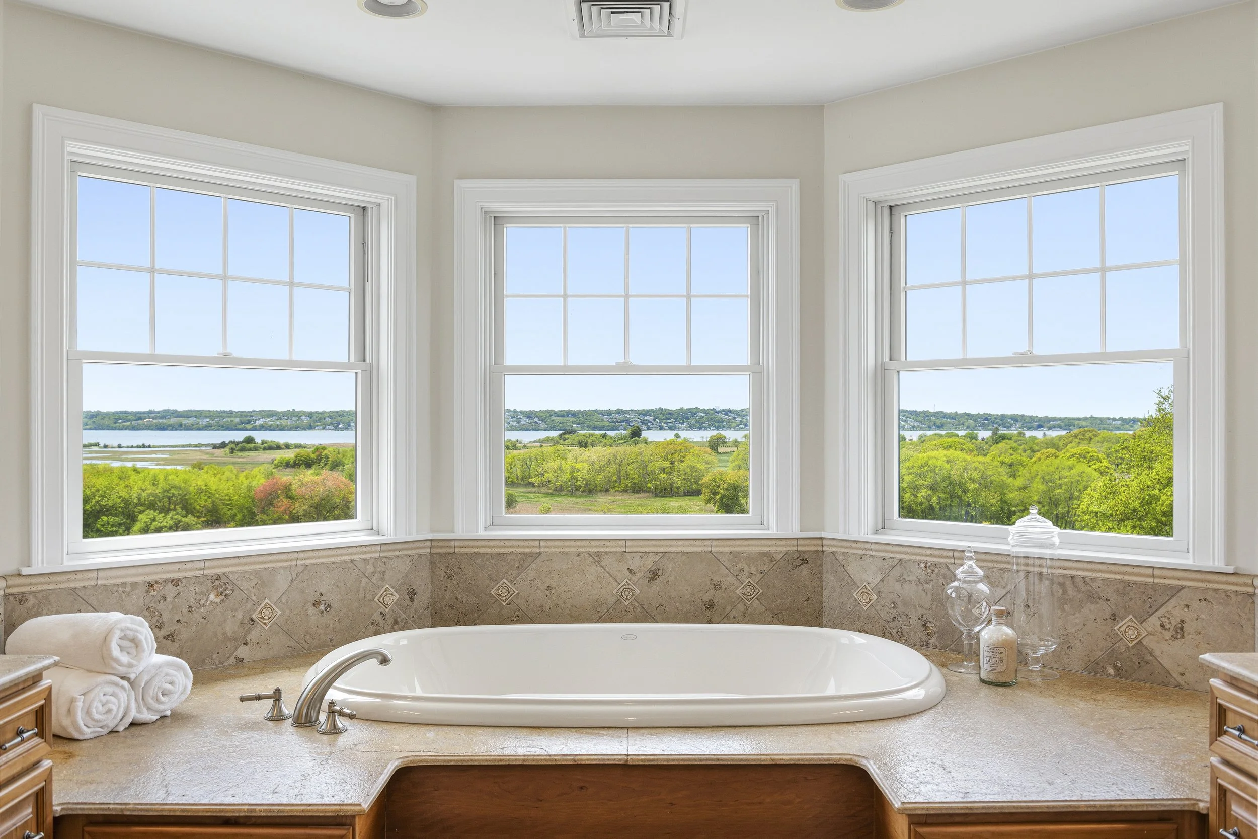A luxurious bathroom with a large bathtub facing three big windows that offer a scenic view of green trees, a river, and a blue sky. The bathroom has beige tile walls, a beige countertop, and wooden cabinetry. There are rolled white towels on the cou