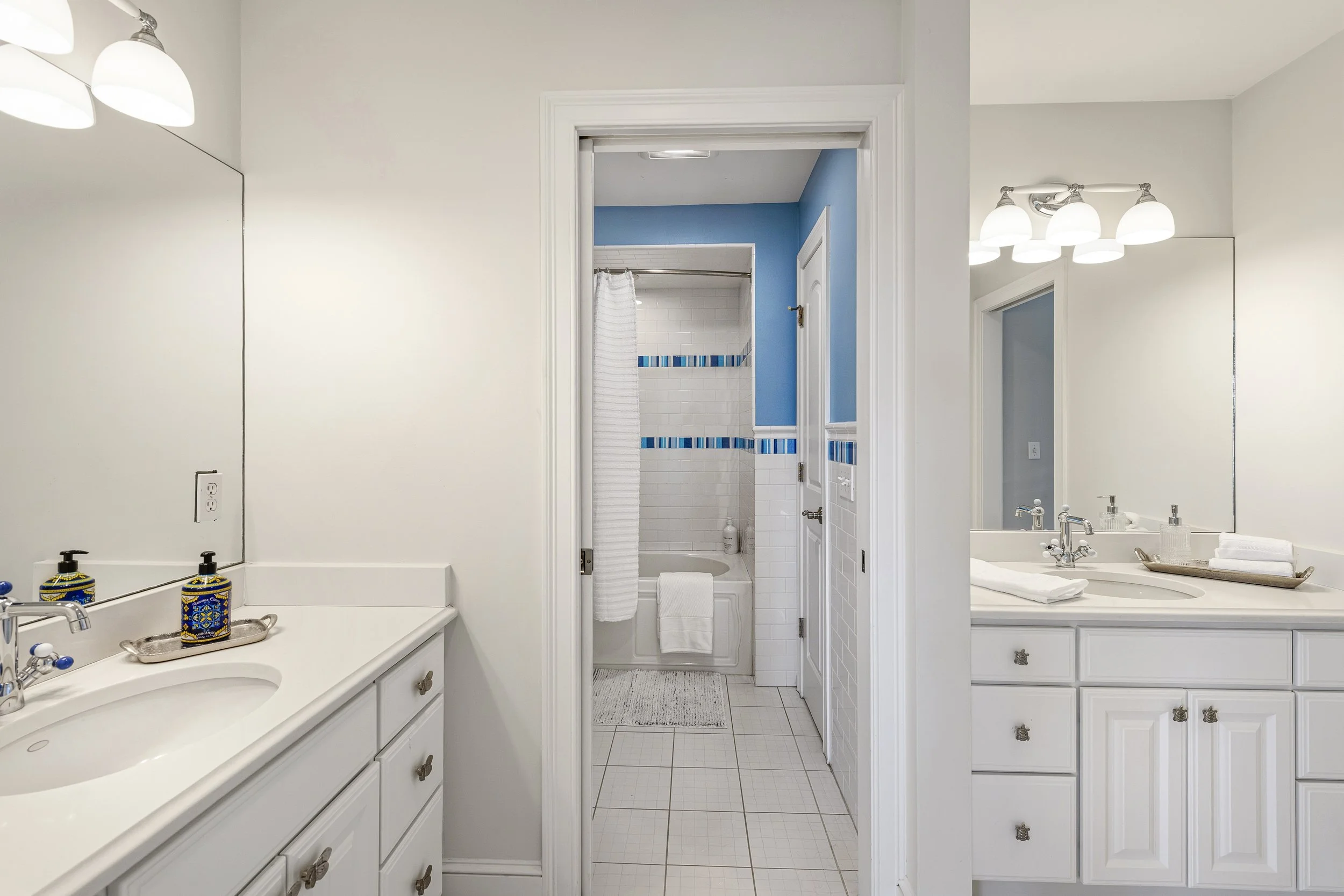 A clean, white bathroom featuring a double vanity with two sinks, chrome fixtures, and neatly folded towels on the countertop. To the left, a mirror reflects part of the room, showing soap dispensers. In the center, there is a doorway with a white do