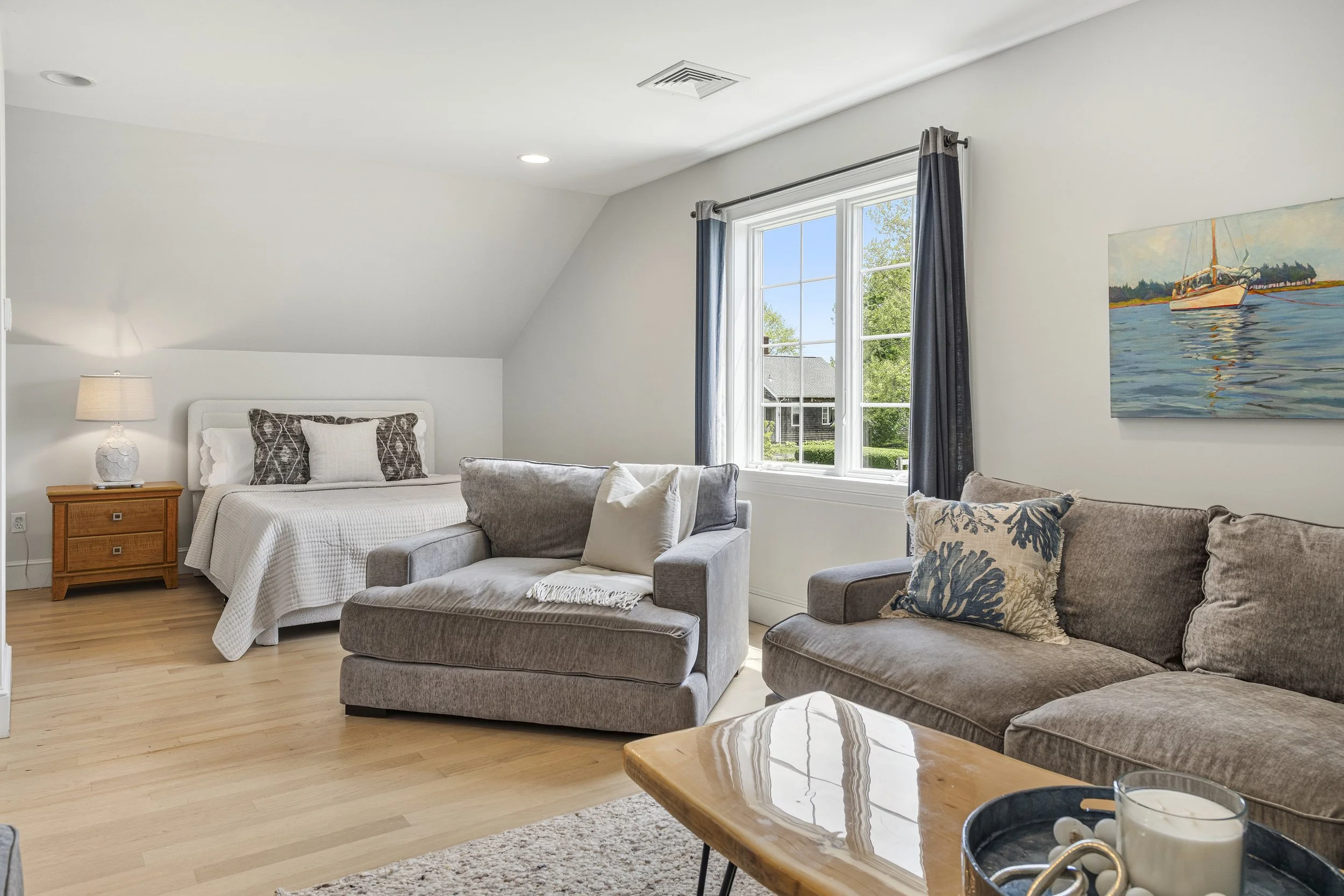 Living room with white walls, large window with blue curtains, tan hardwood floors, white bed with pillows, gray couches, painting of sailboats on the water, wooden coffee table with candles and decorative items.