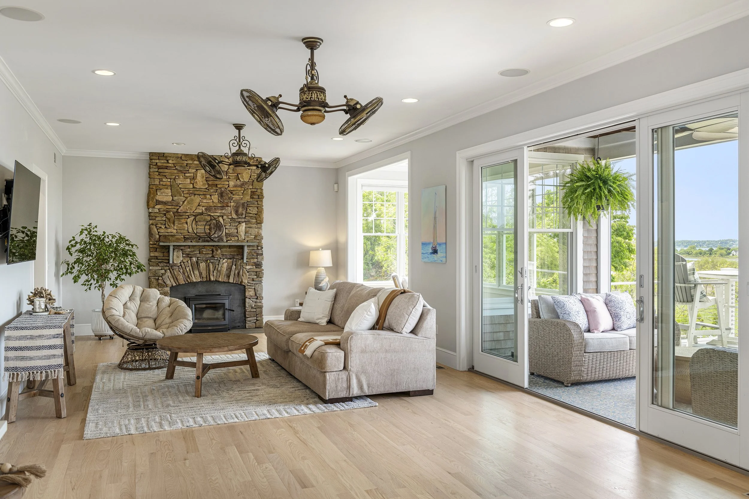 Living room with beige couch and armchair, stone fireplace, large windows, and sliding glass door leading to an outdoor patio with greenery.