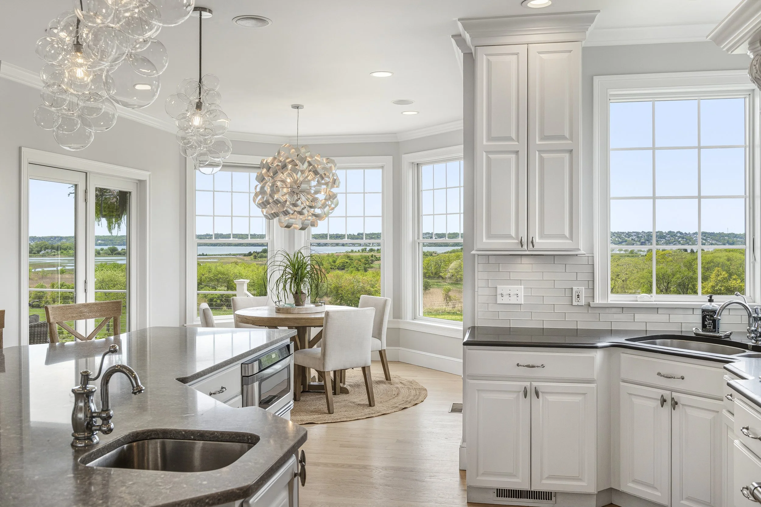 Bright Kitchen with large windows overlooking green landscape, white cabinetry, gray countertops, modern light fixtures, and a round dining table with chairs.
