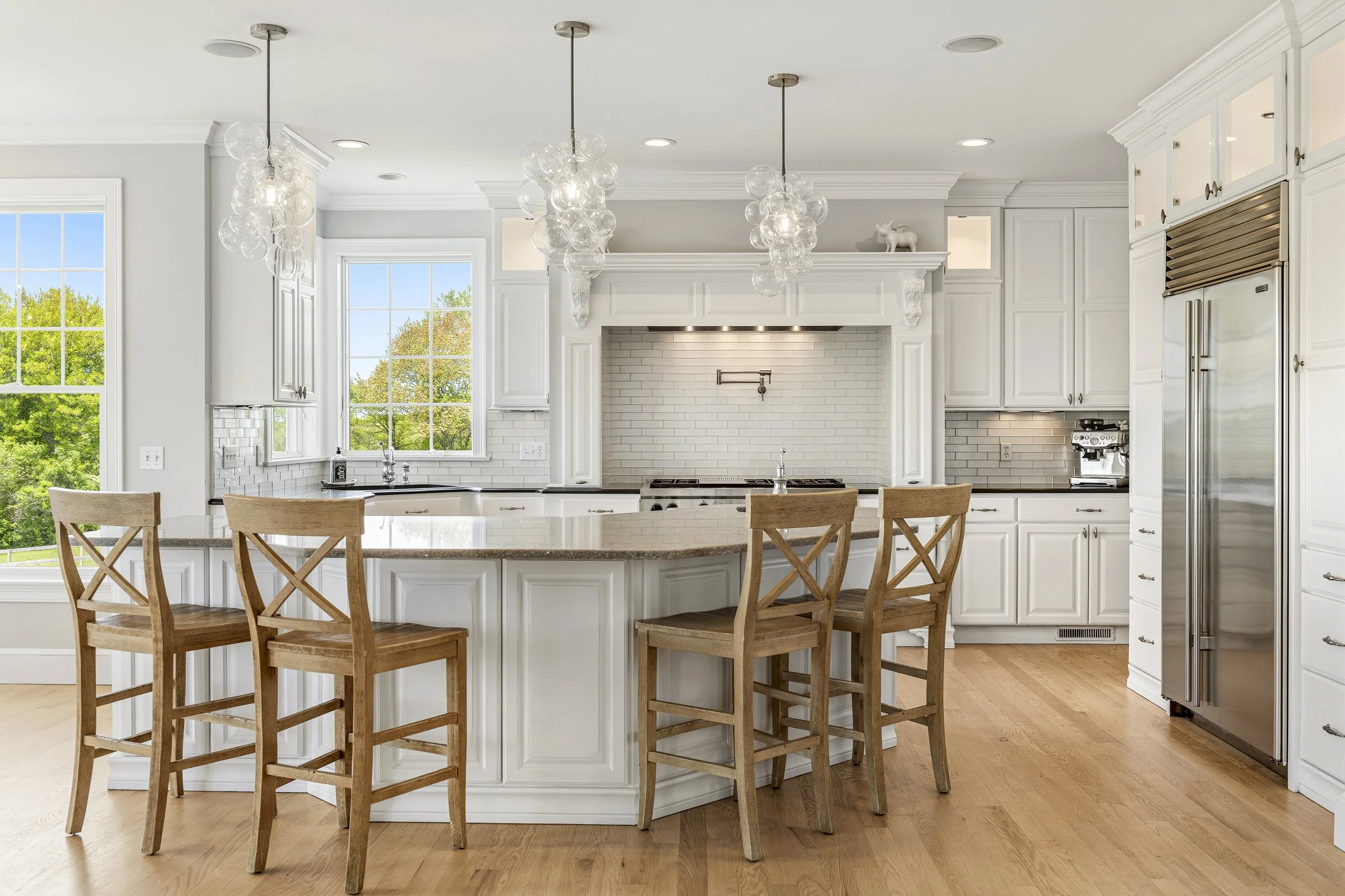 Modern kitchen with white cabinets, large island with wooden chairs, brick backsplash, stainless steel appliances, and multiple large windows showing green trees outside.