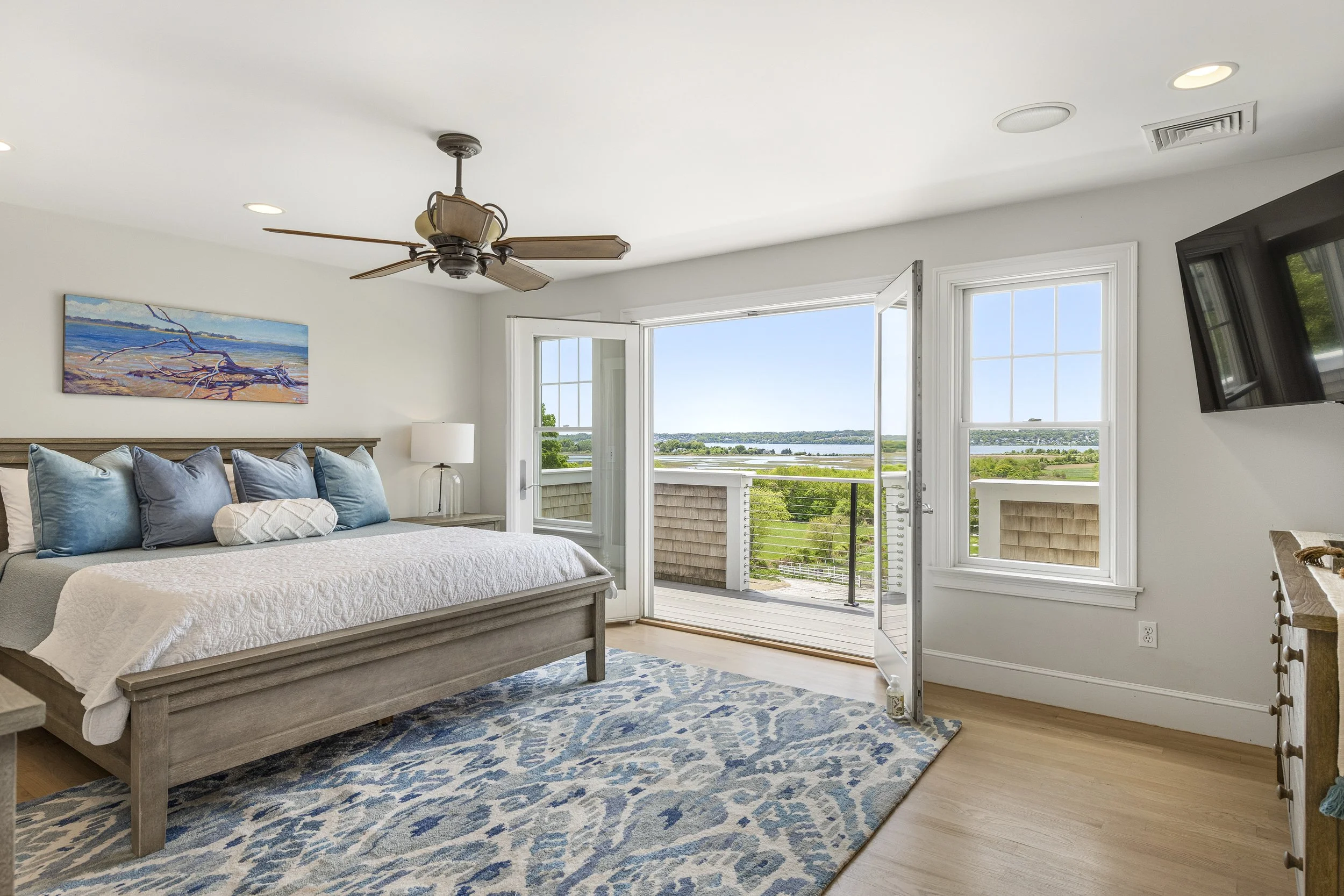 A bedroom with a large window showing a view of green landscape and water, a bed with blue and white pillows, a painting of a beach scene on the wall, a ceiling fan, and a door leading to a balcony.